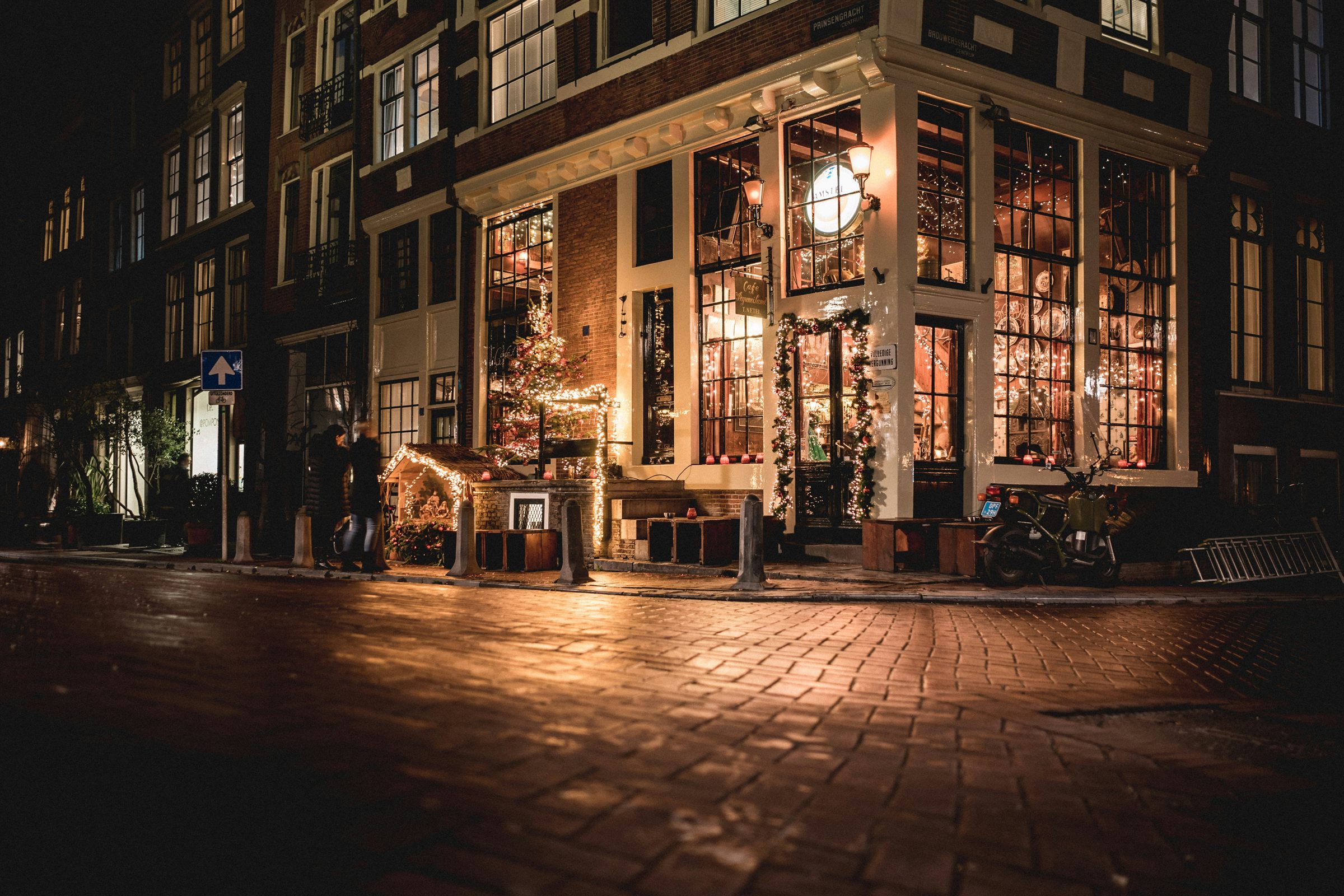 A corner cafe lit up at night, its windows glowing over wet cobblestones