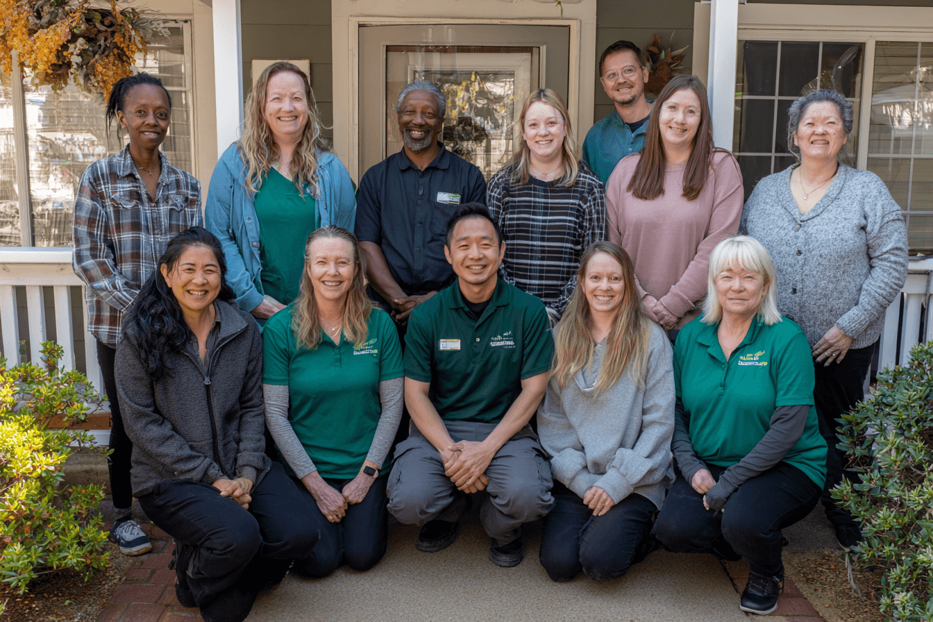 a home care agency. Caregivers are gathered around the home care agency operator, who is wearing a dark green shirt. They are all smiling and posing for a picture