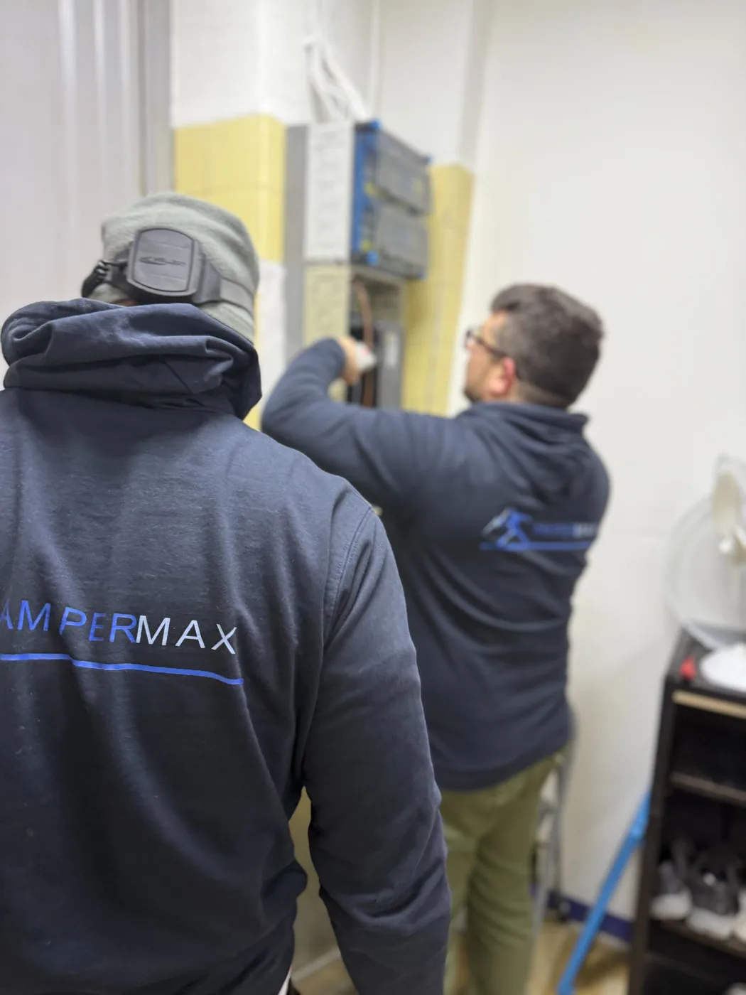 Két Ampermax villanyszerelő közösen dolgozik egy elosztótáblán Ampermax pulóverben