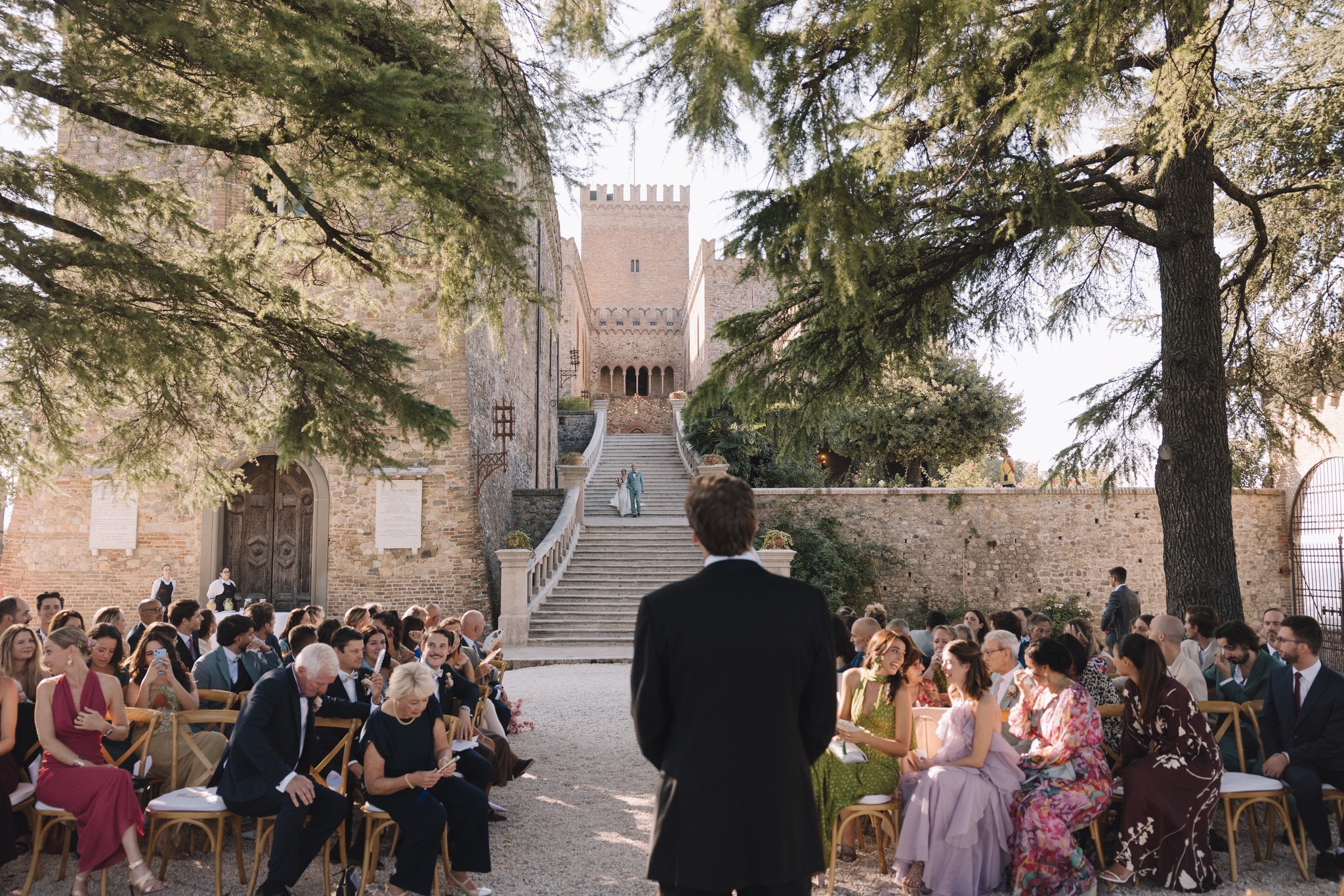Cerimonia di matrimonio al Castello di Tabiano con sposi e ospiti sotto gli alberi, scalinata della Rocca sullo sfondo