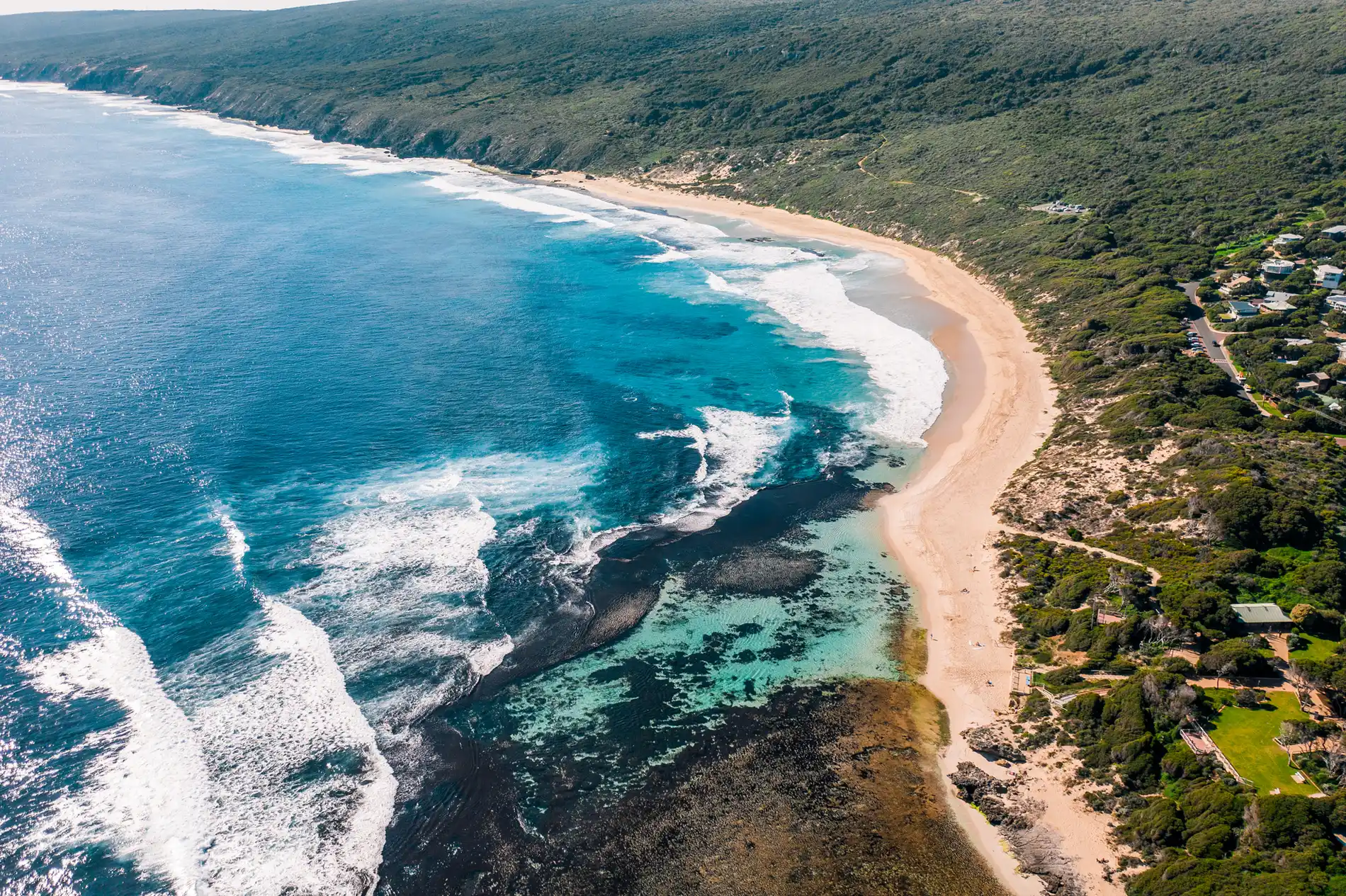 Aerial of a long surf beach curving beneath green coastal bushland