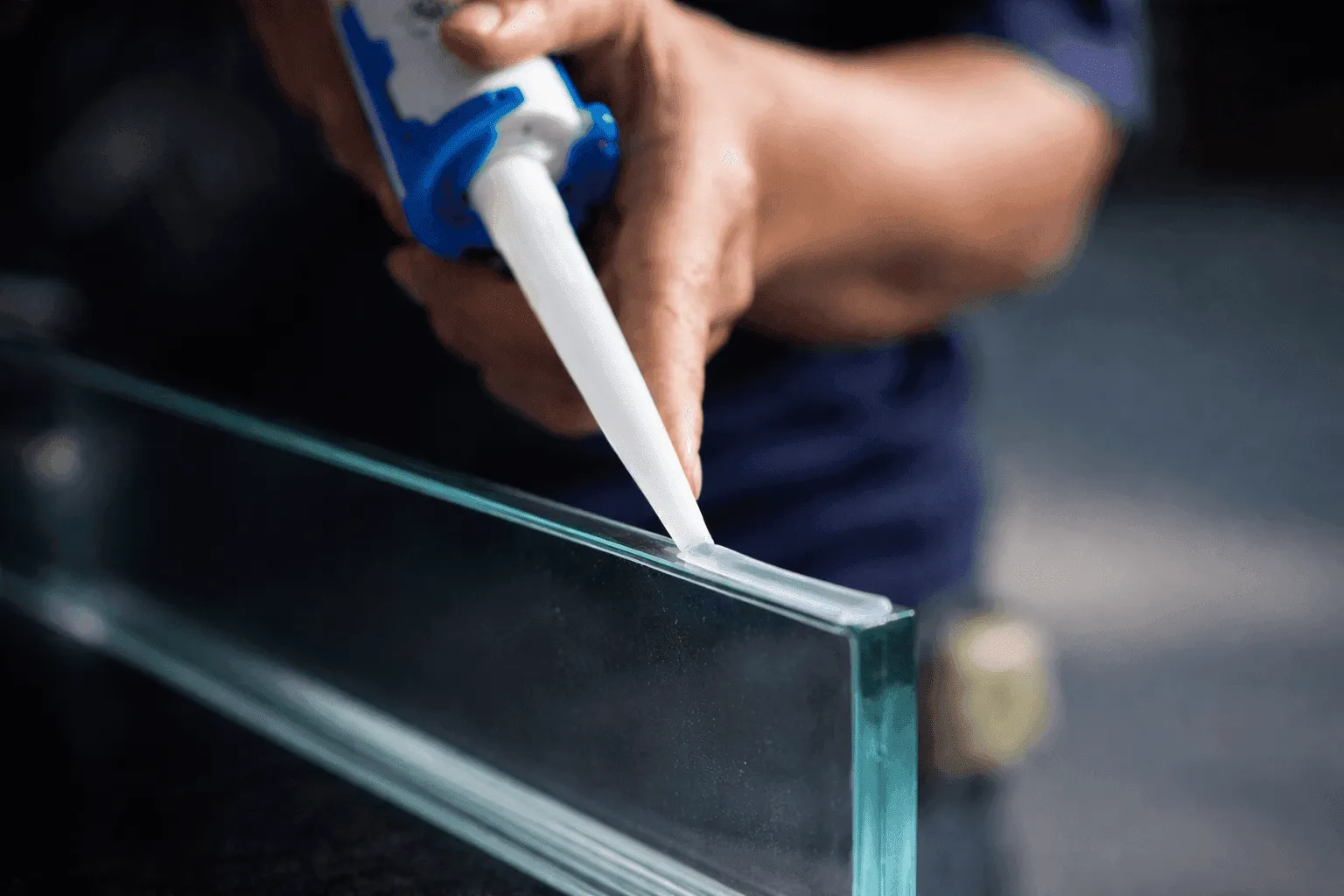 A close-up of a hand applying sealant to the edge of a glass surface with a caulking gun.
