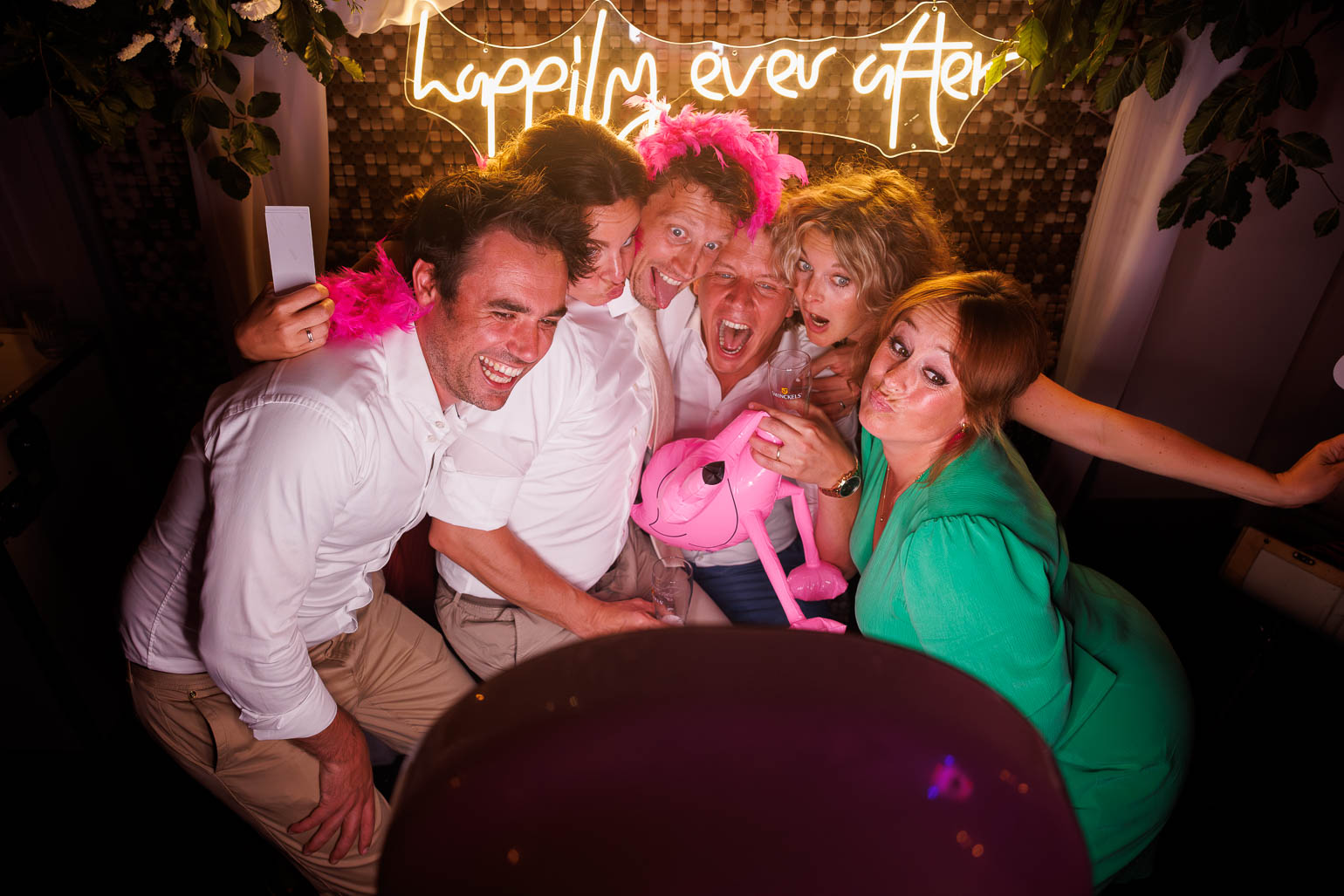 Wedding guests are posing for photos with a flamingo inflatable. Neon-lit background: Happily ever after.