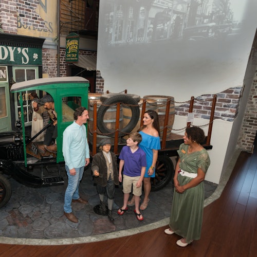 三位成人和两位儿童站在一个历史展览的旧绿色送货卡车旁,周围是砖墙和复古装饰。