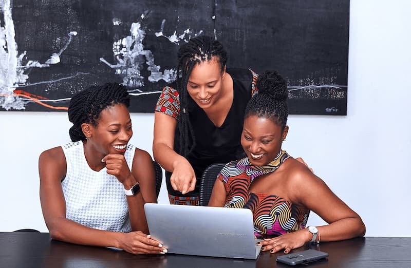 Trois femmes souriantes regardent un ordinateur portable autour d’une table. - Women in Afrofood