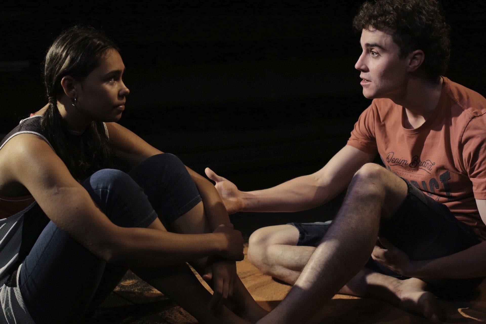 Two young performers sit barefoot on a stage covered with reddish sand, facing each other. The girl listens intently with her knees pulled close to her chest, while the boy leans forward, gesturing as he speaks. Dramatic lighting highlights their serious expressions.