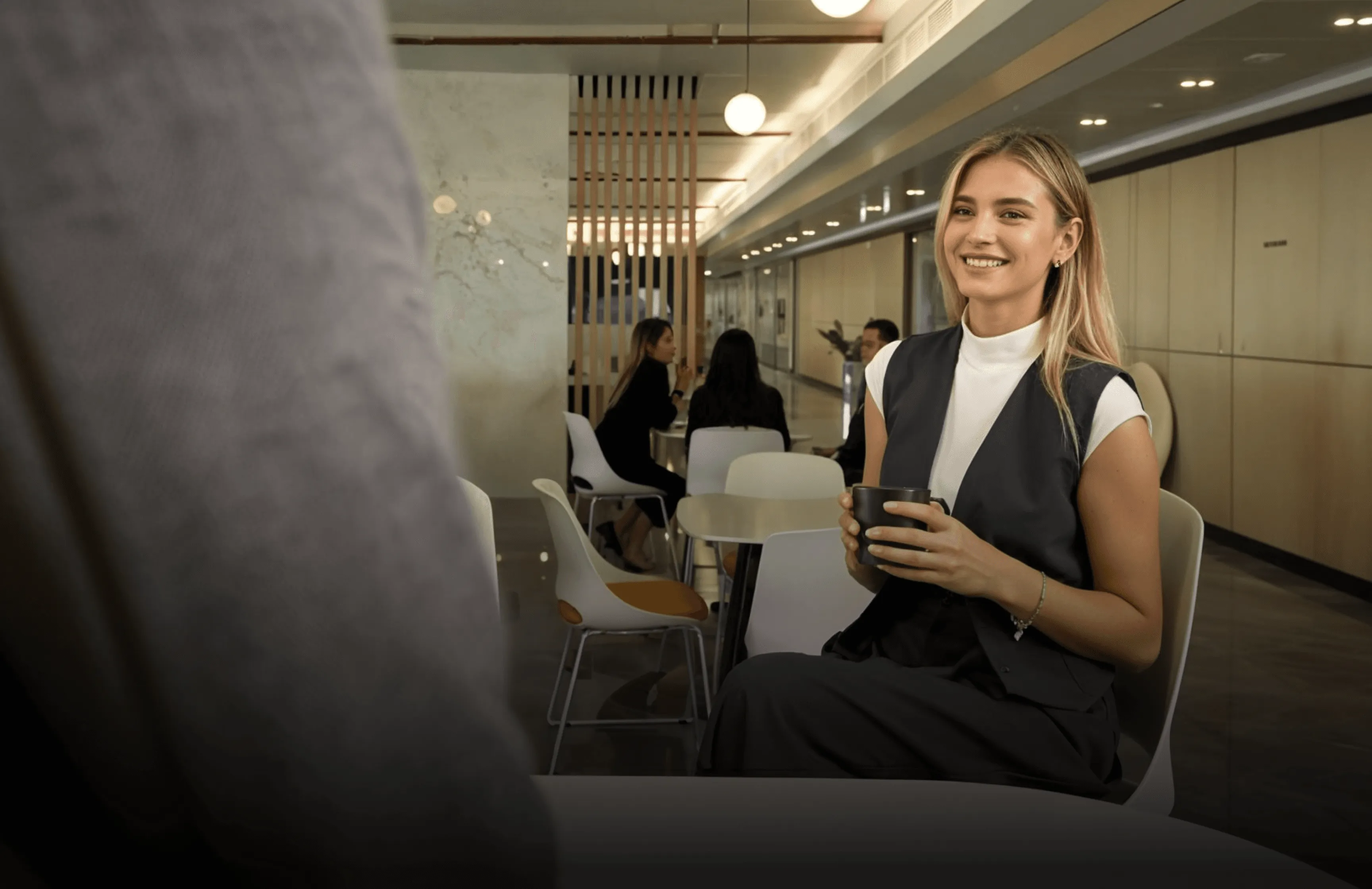 Blonde woman in a vest holding a coffee cup, smiling. Other people seated at tables, interior