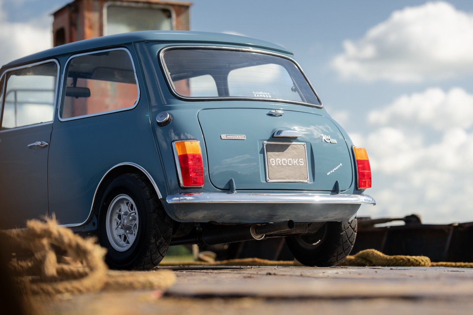 A blue vintage car parked on a sandy surface, with a scenic background of clouds and sky.