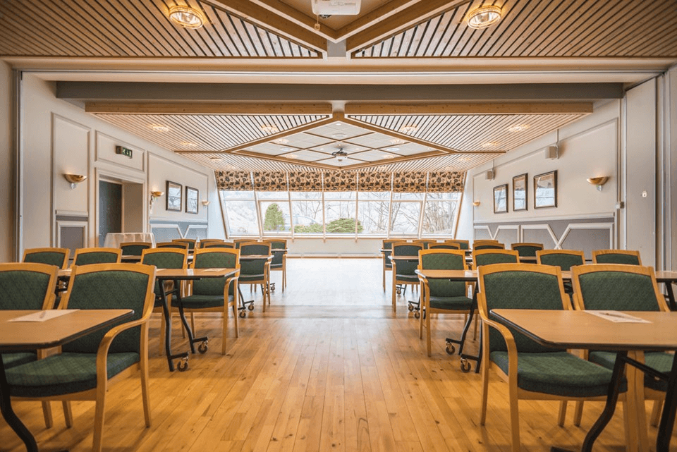 Brightly lit dining area with wooden floors and green chairs arranged around tables, leading to an outdoor view.