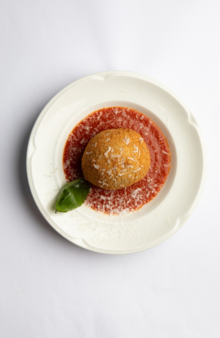 Une boule d'arancini dorée et croustillante repose sur un lit de sauce tomate rouge riche, garnie de basilic frais et saupoudrée de fromage râpé, servie sur une assiette blanche, contre un fond uni.
