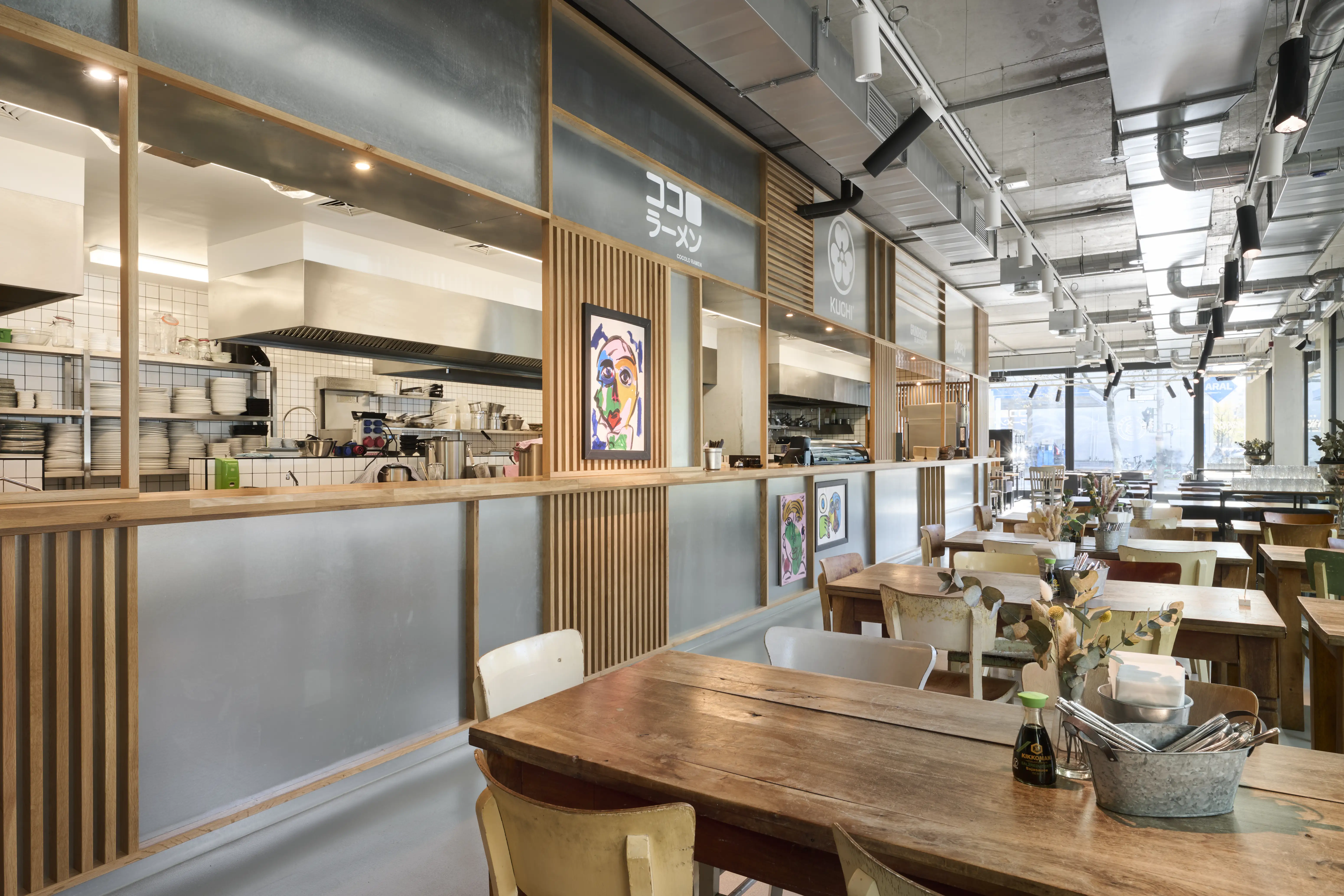 Dining room at PARKER BOWLES & FRIENDS with rustic wooden tables, mixed vintage chairs, a wood-and-frosted-glass kitchen front with Japanese ramen signage and colorful artwork.