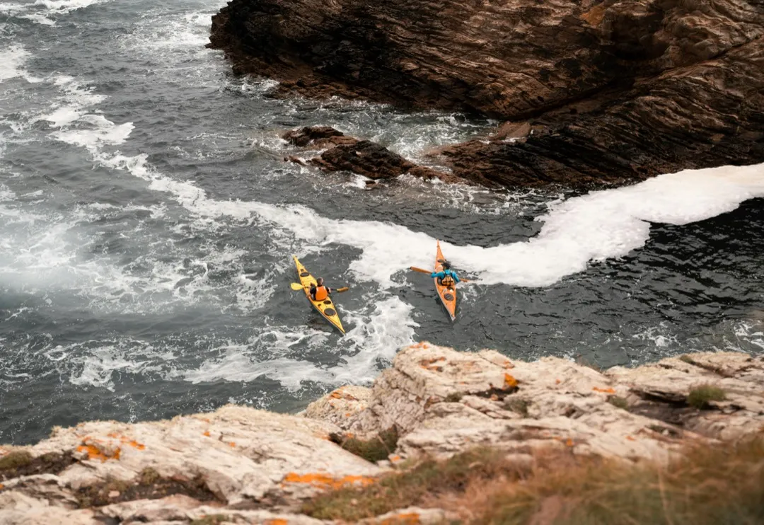 two people in separate kayaks navigate a rocky shoreline