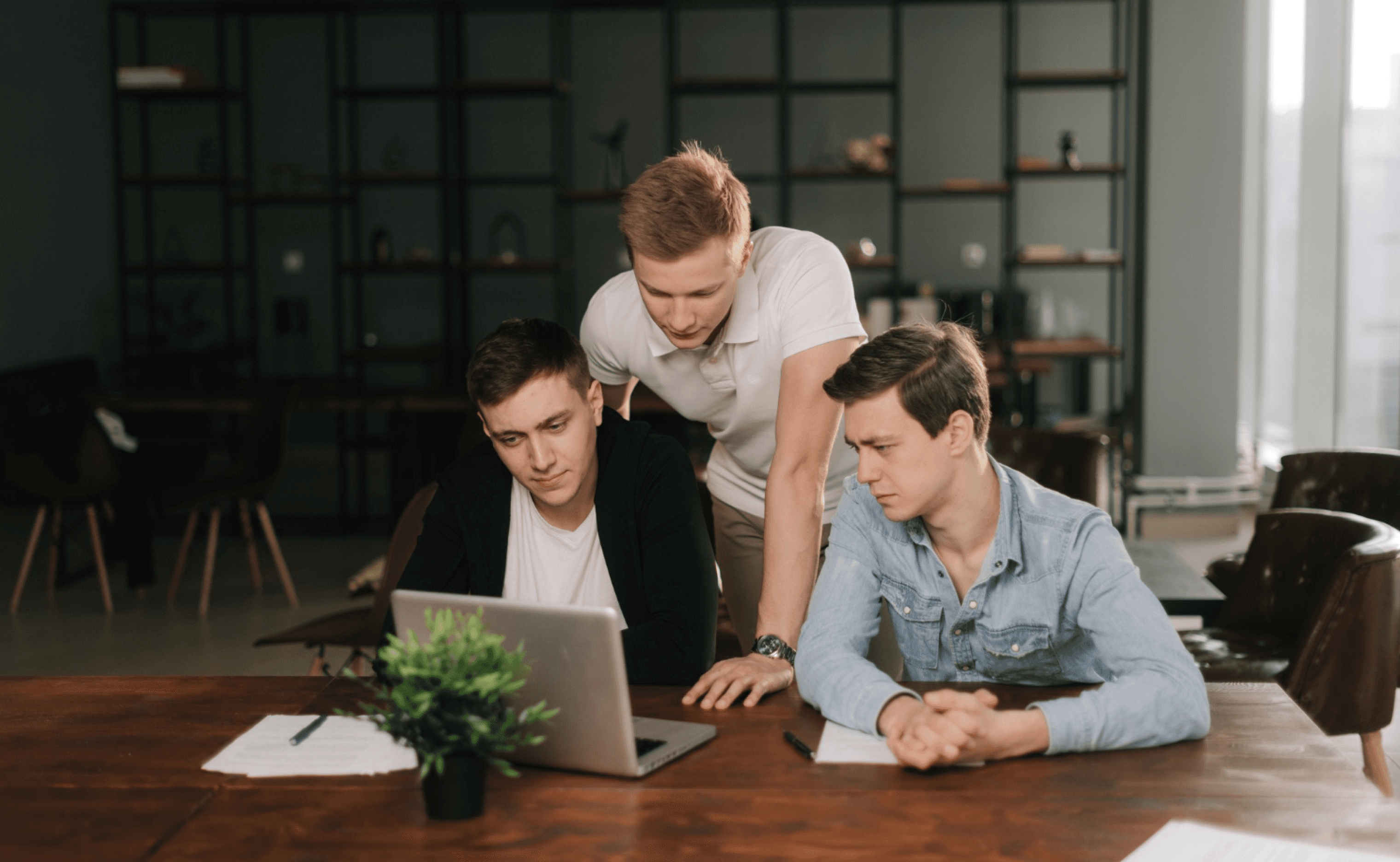 Three people collaborating around a laptop at a table with papers and a plant