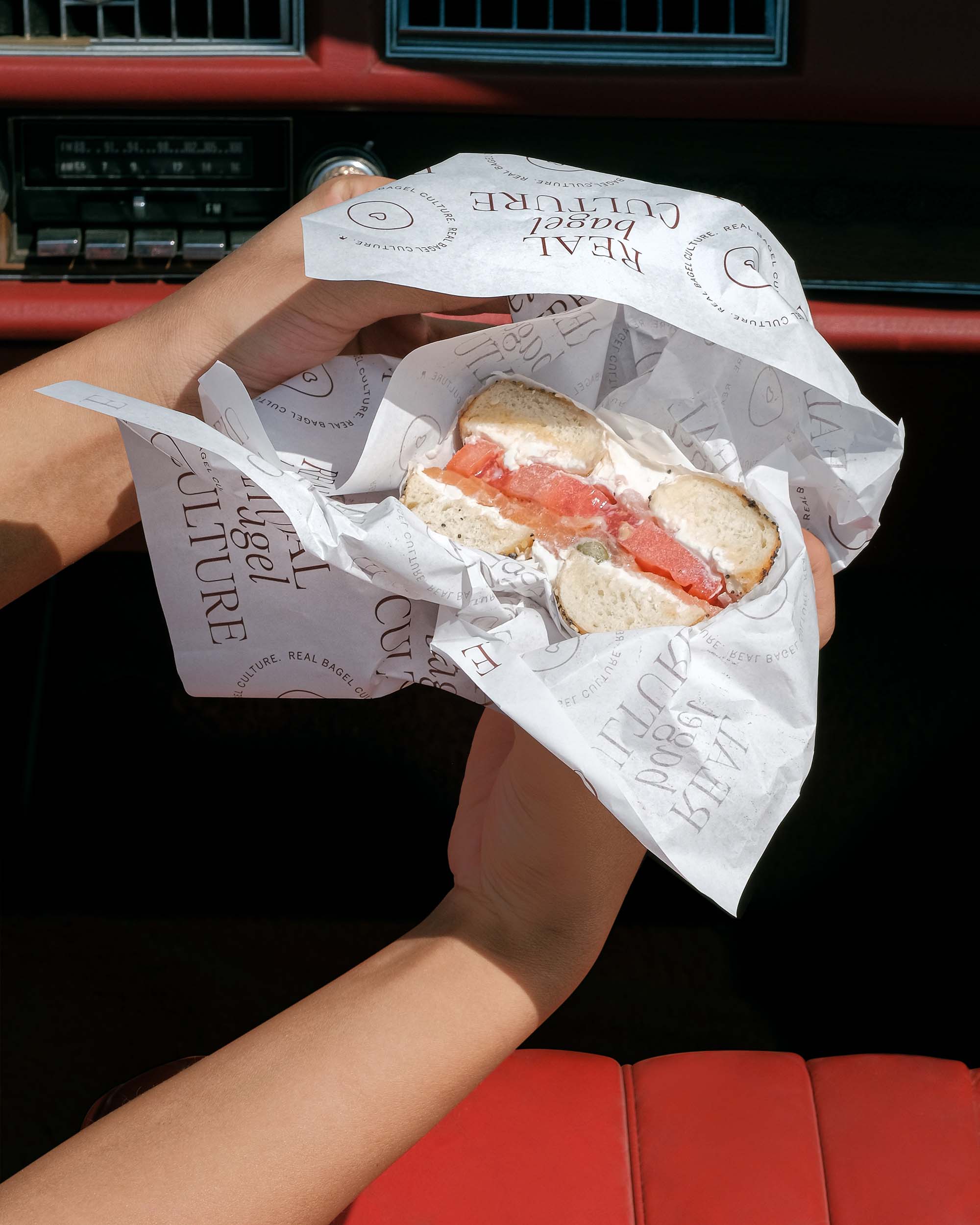 Person holding a bagel sandwich with cream cheese and salmon wrapped in paper. Text on the paper says "REAL BAGEL CULTURE."