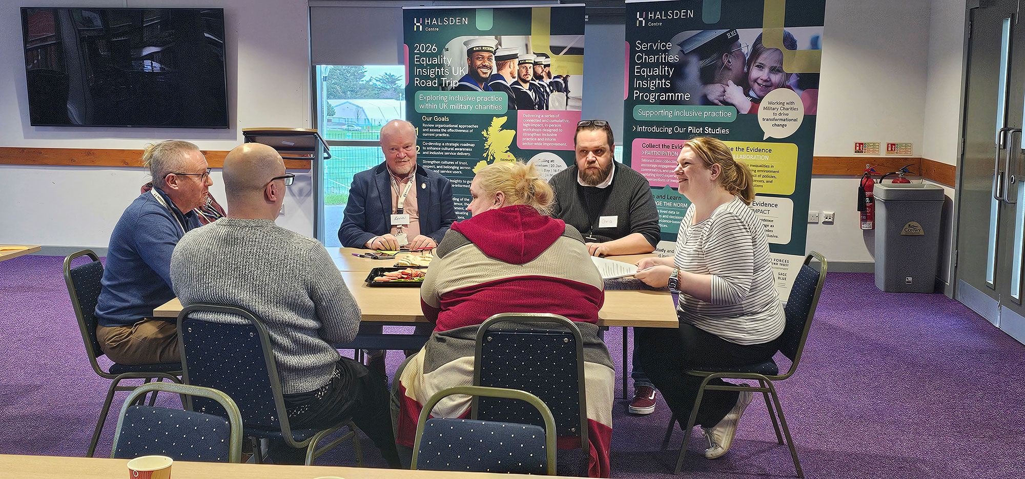 A group of people seated around a table in discussion at a Halsden Centre event, with branded roll-up banners for the Service Charities Equality Insights Programme in the background.