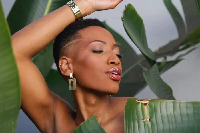 A woman with closed eyes and glossy lips, one arm raised over her head, framed by broad tropical leaves