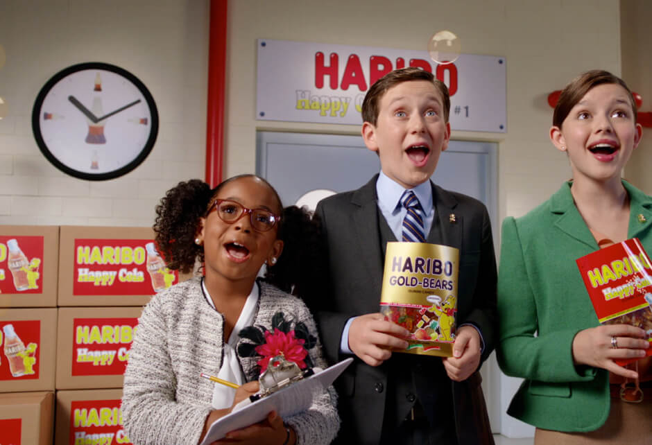 Three children dressed as adults stand in an office setting, holding Haribo candy products, with boxes and a clock in the background.
