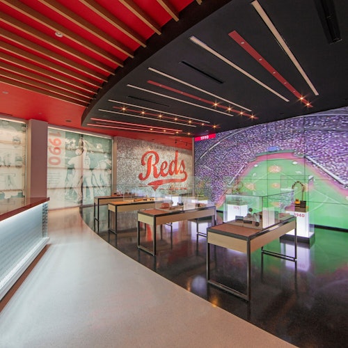 Modern museum exhibit featuring baseball memorabilia, a large "Reds" sign, and a giant screen displaying a crowded stadium.