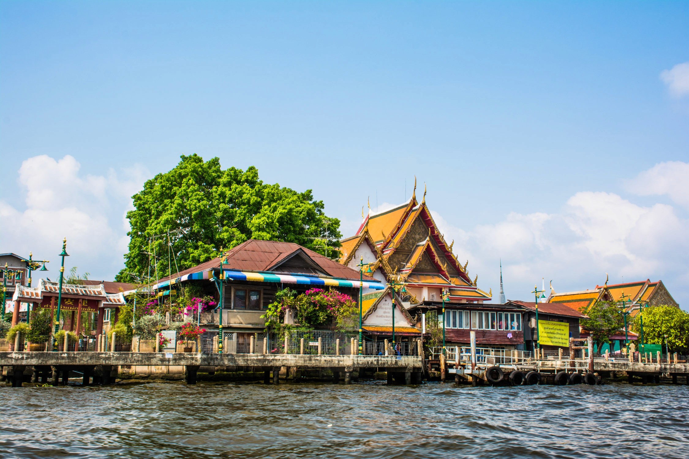 A Thai temple complex with golden roofs and colorful buildings sits along a waterfront under a clear blue sky.