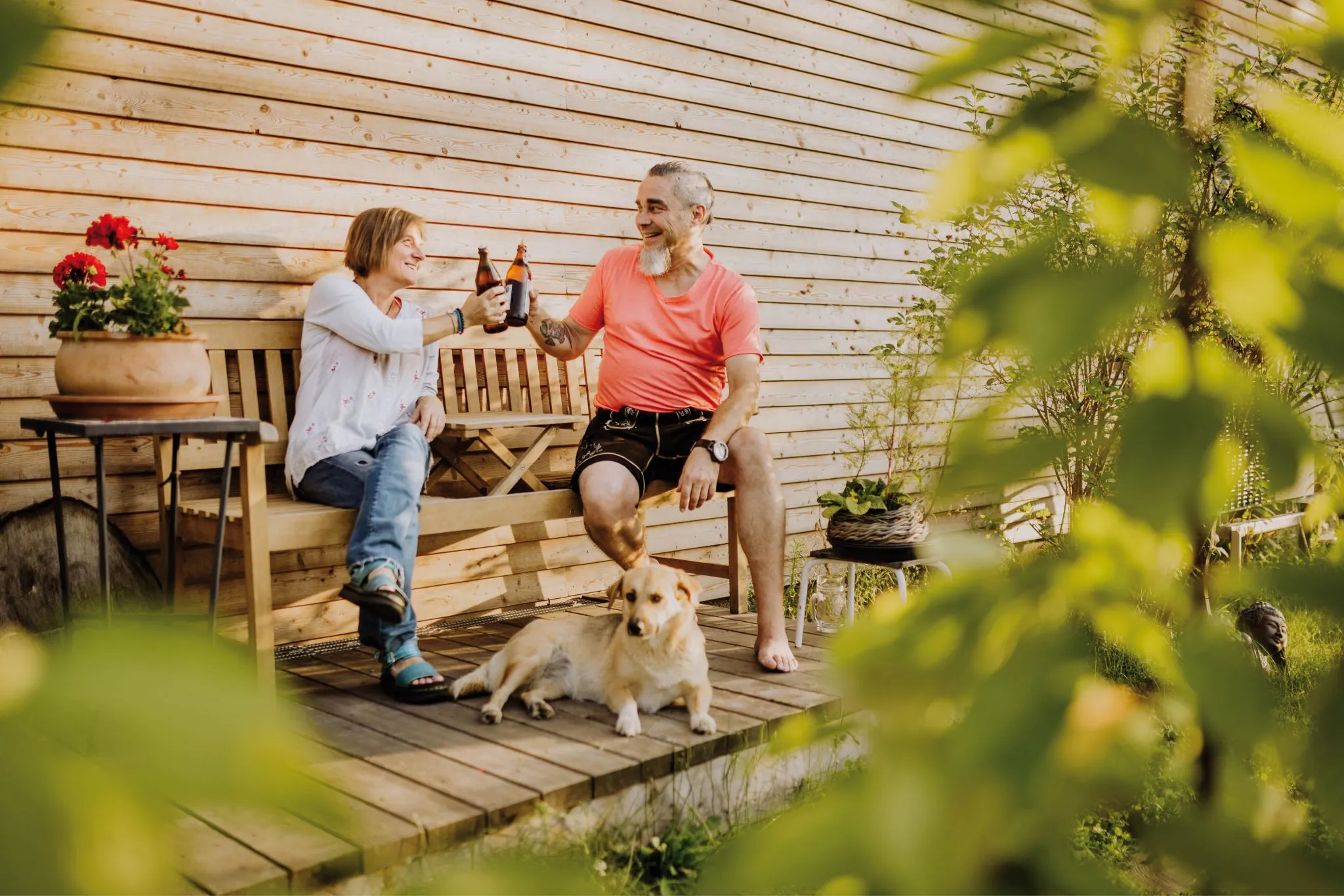 Ein Paar stößt mit Bierflaschen an, während ihr Hund vor einer Holzhütte entspannt auf der Terrasse liegt.
