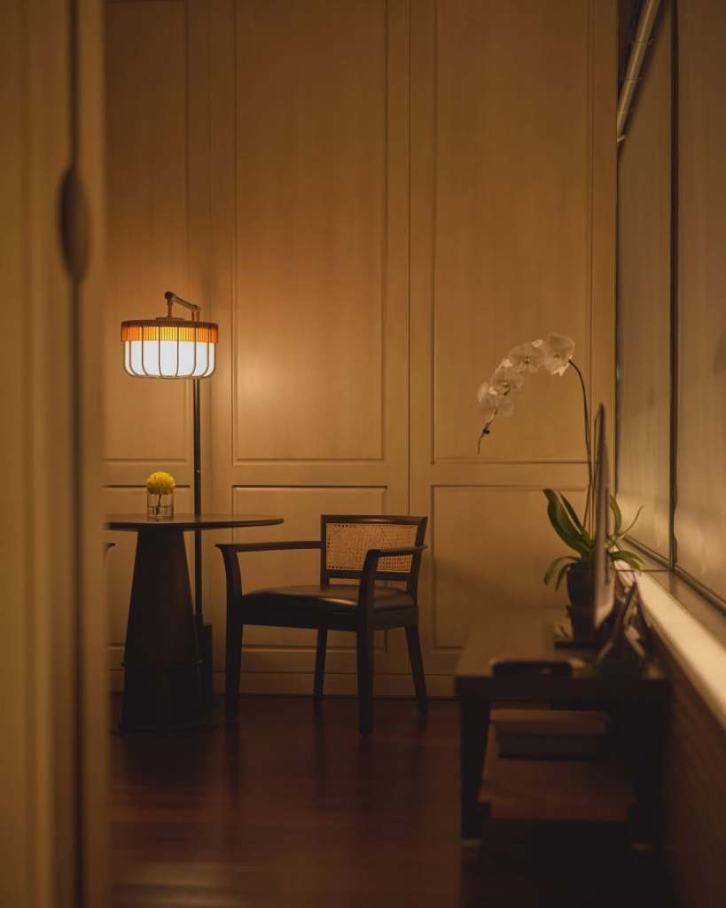 A cozy corner in a softly lit room featuring a small round table with a yellow flower in a glass, a wooden chair, and a floor lamp with a white and orange shade against a backdrop of wooden paneling.