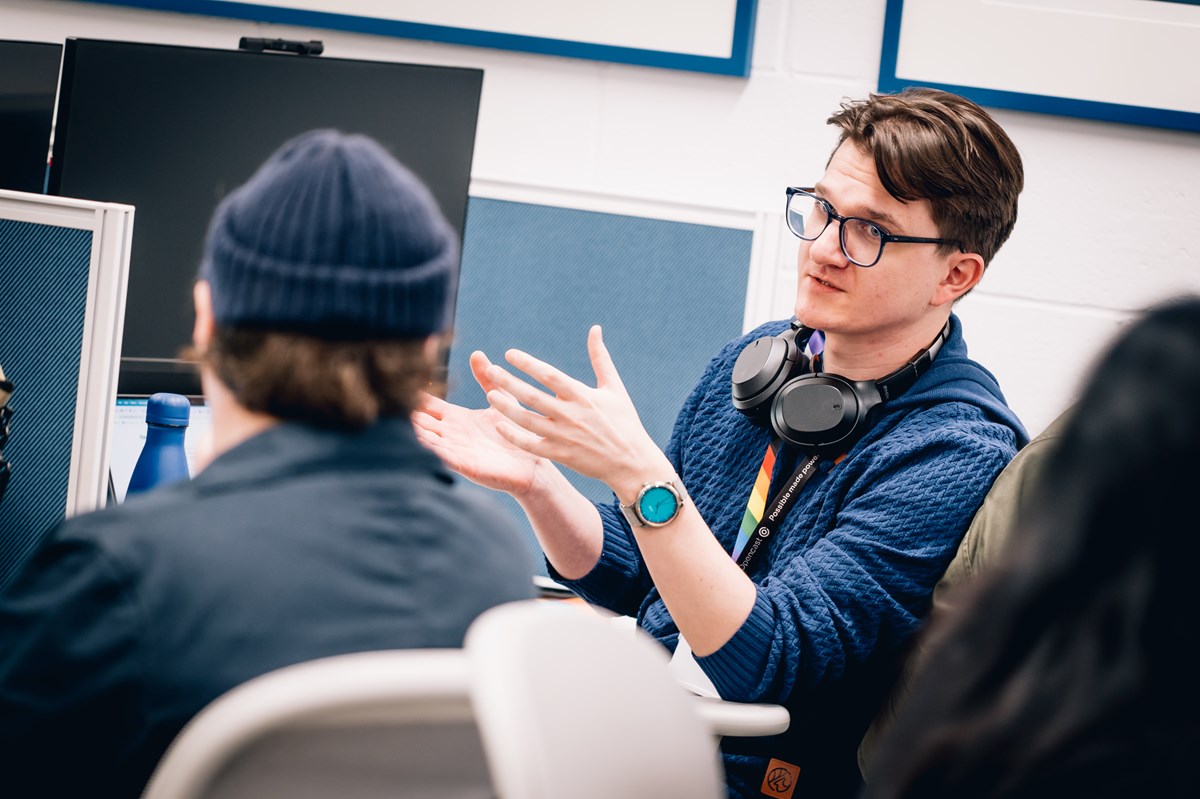 A man in a blue shirt wearing glasses and headphones round his neck gestures while conversing with a person wearing a blue hat out of focus in the foreground, they are sat at desks with computer monitors on them