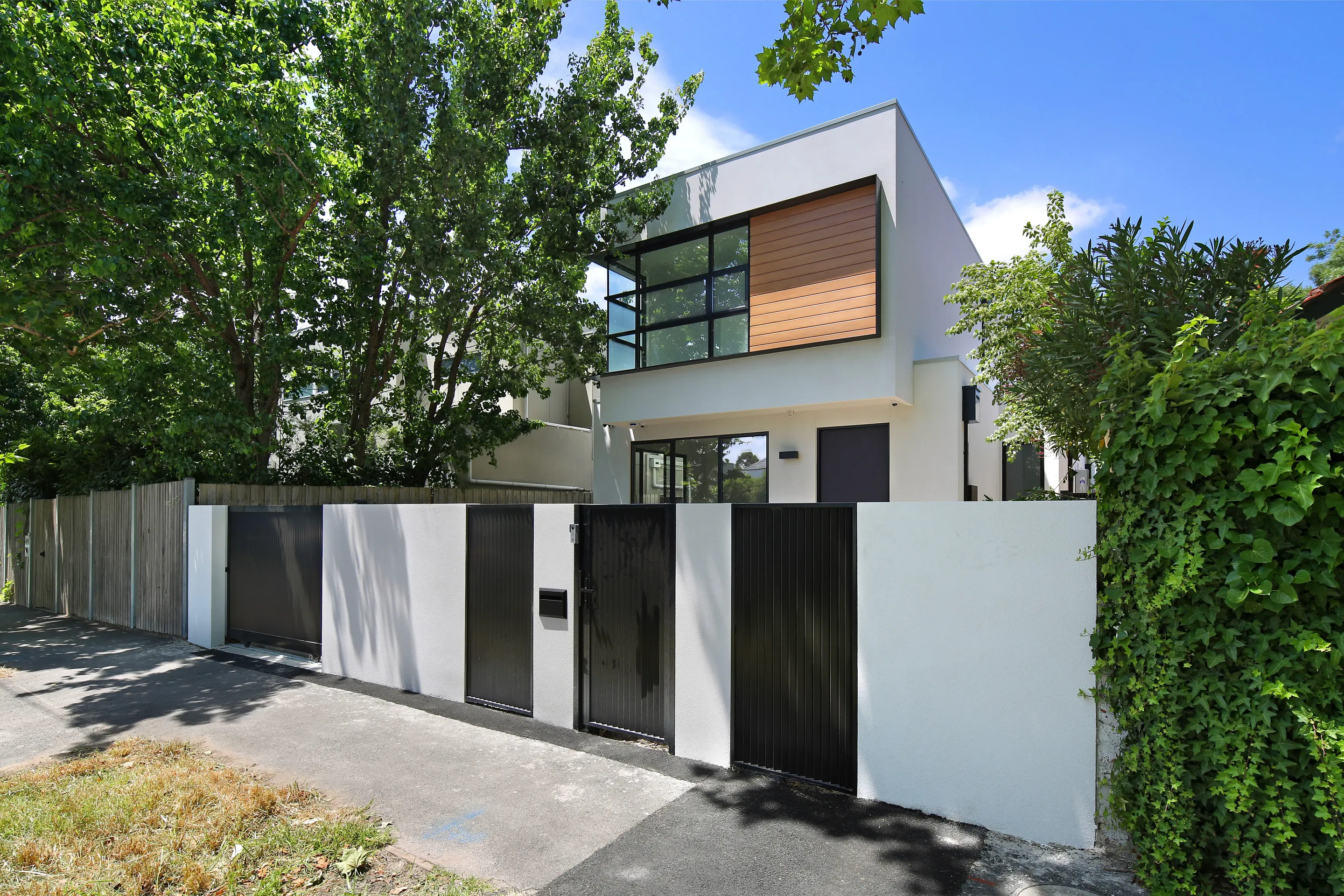 Street view of the Malvern Residence, a crisp white double-storey home with a timber-clad upper box and black-framed windows, behind a rendered front wall with black slatted gates among leafy trees