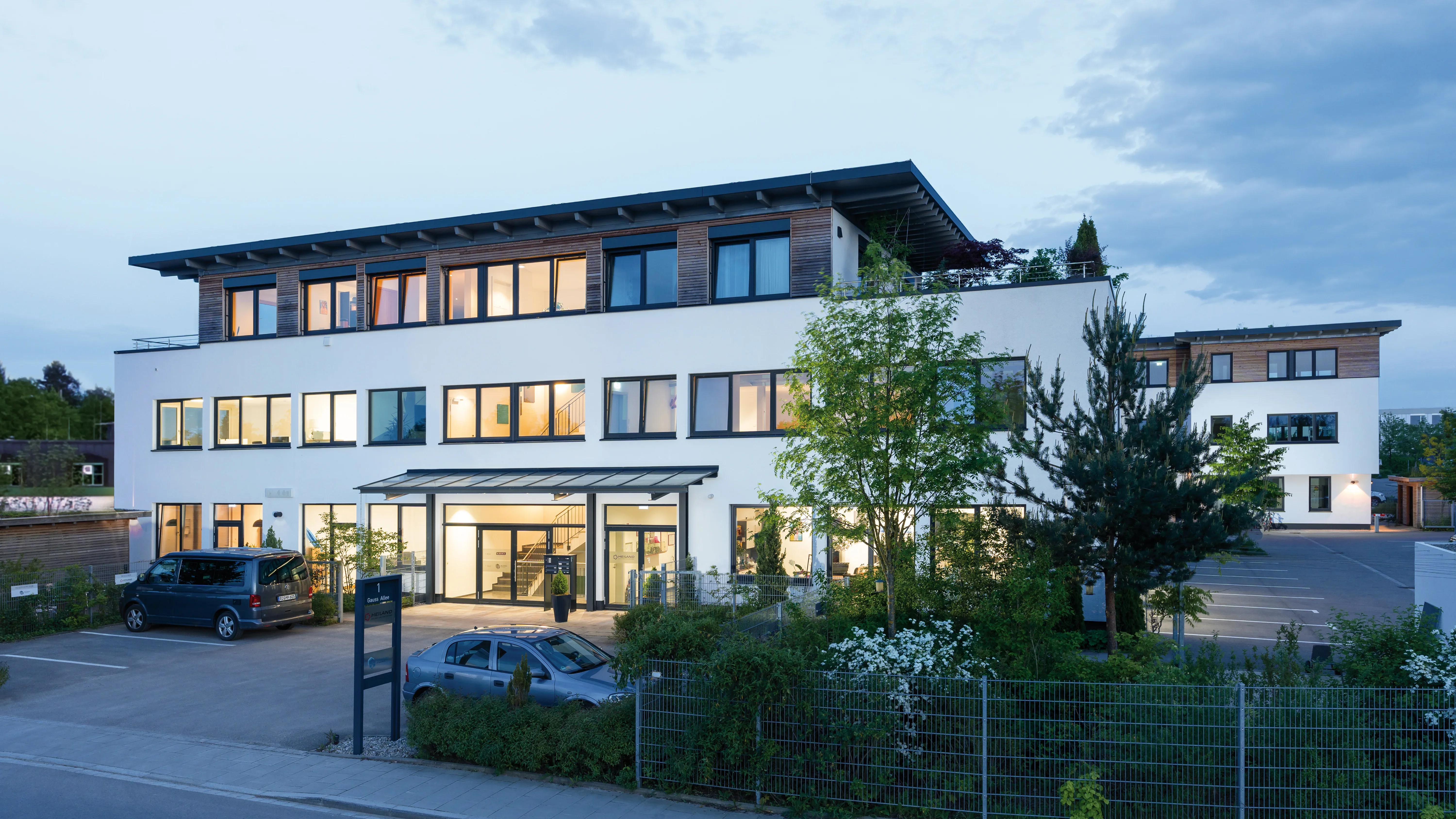 Modernes Bürogebäude mit Holzverkleidung, beleuchteter Eingang und Parkplatz im Abendlicht.