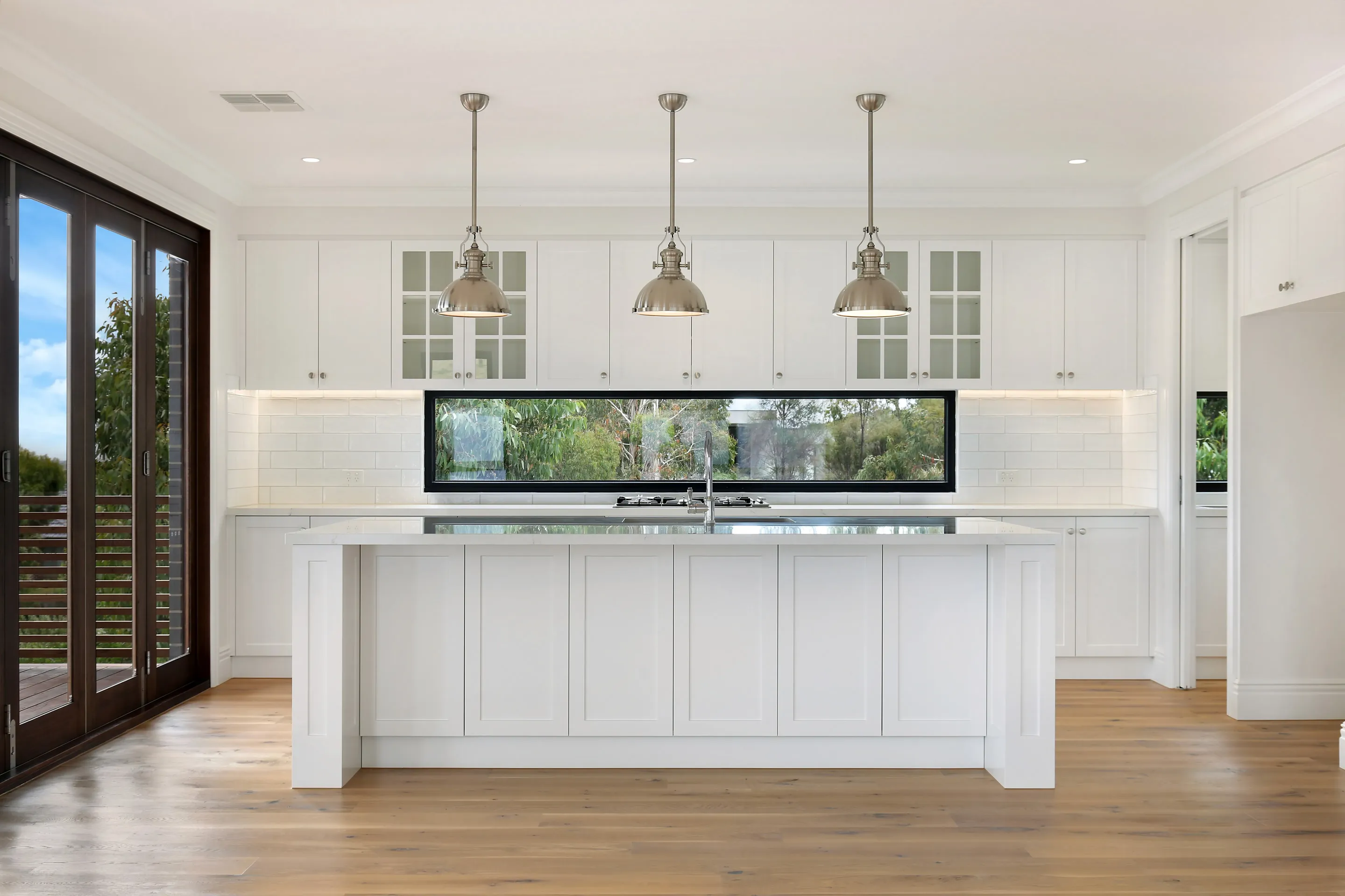 White shaker kitchen viewed head-on with a central island, three polished-nickel dome pendants, a long black-framed window with bush views and a subway-tile splashback