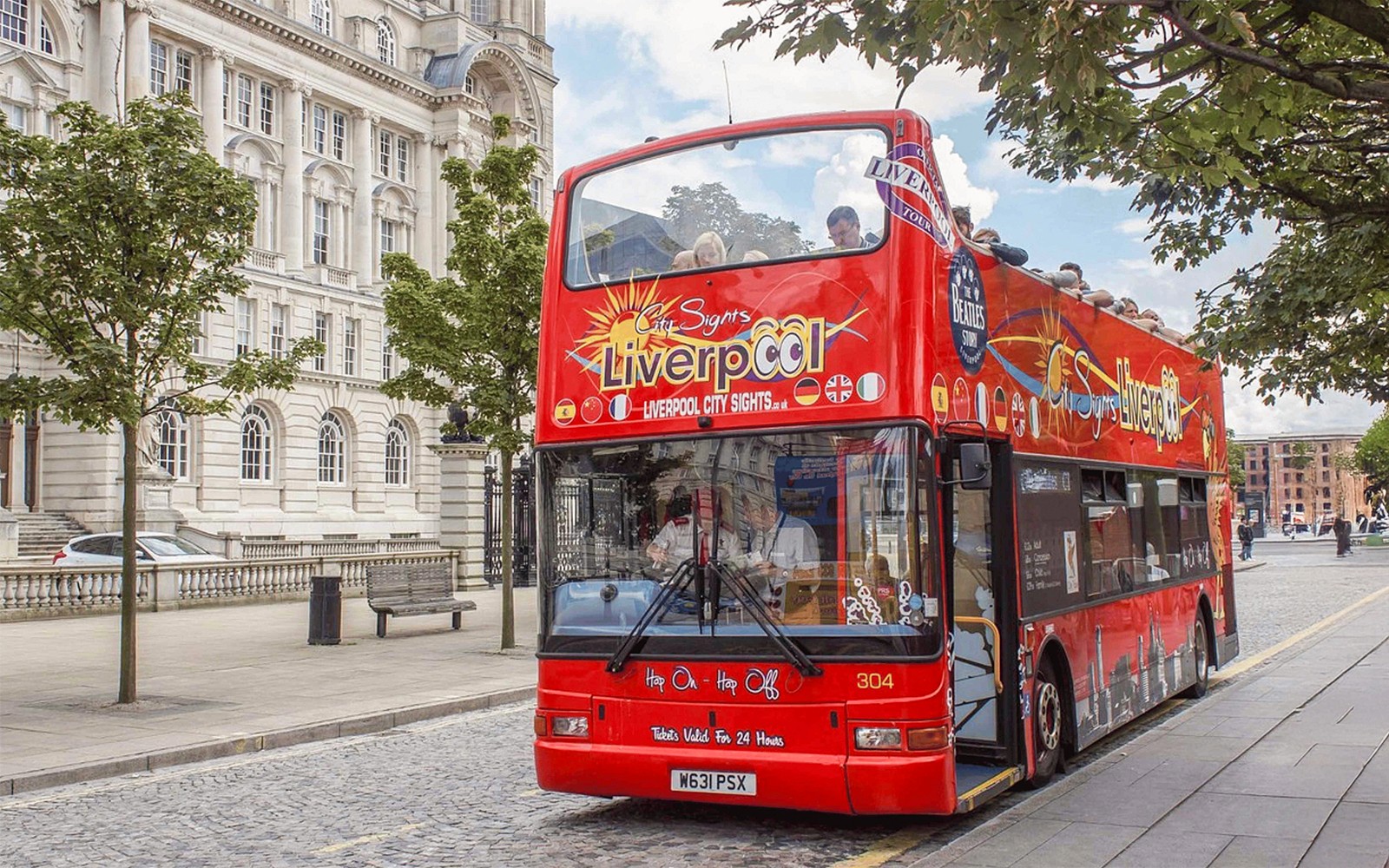 Red double-decker bus on Liverpool Hop On Hop Off City Tour near historic building.