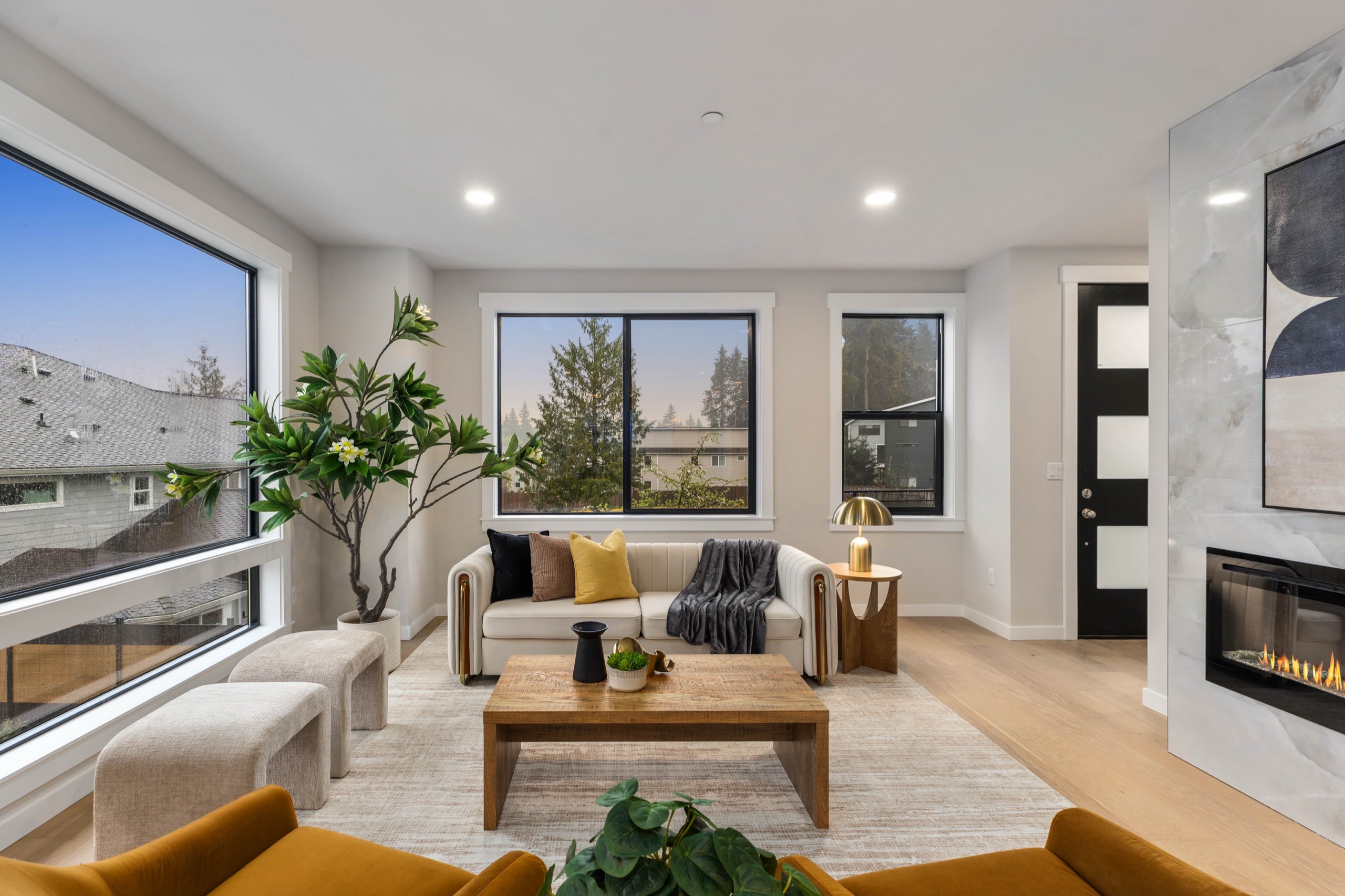 Sun-filled luxury living room with oversized picture windows, neutral sectional sofa, wood coffee table, hardwood floors, and modern linear fireplace with white surround