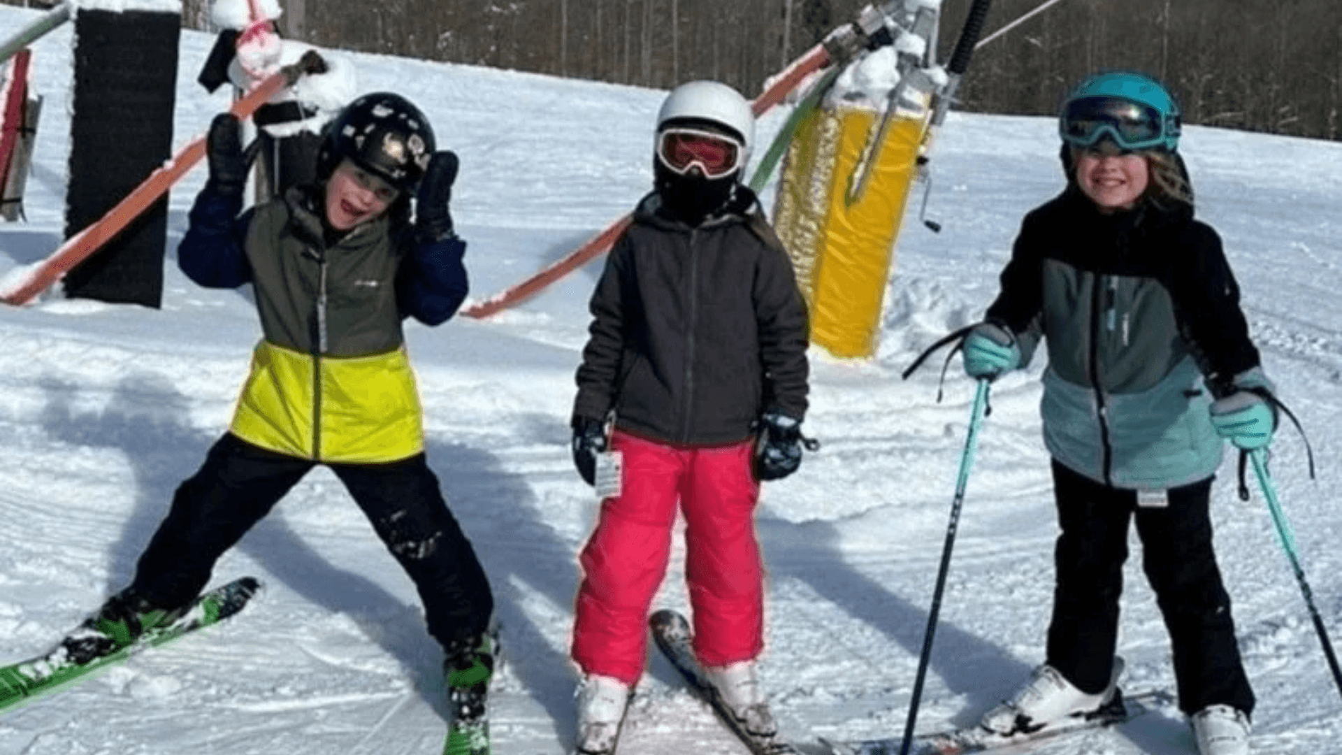 Alt tag: Three children wearing helmets and winter gear smiling while standing on skis in the snow