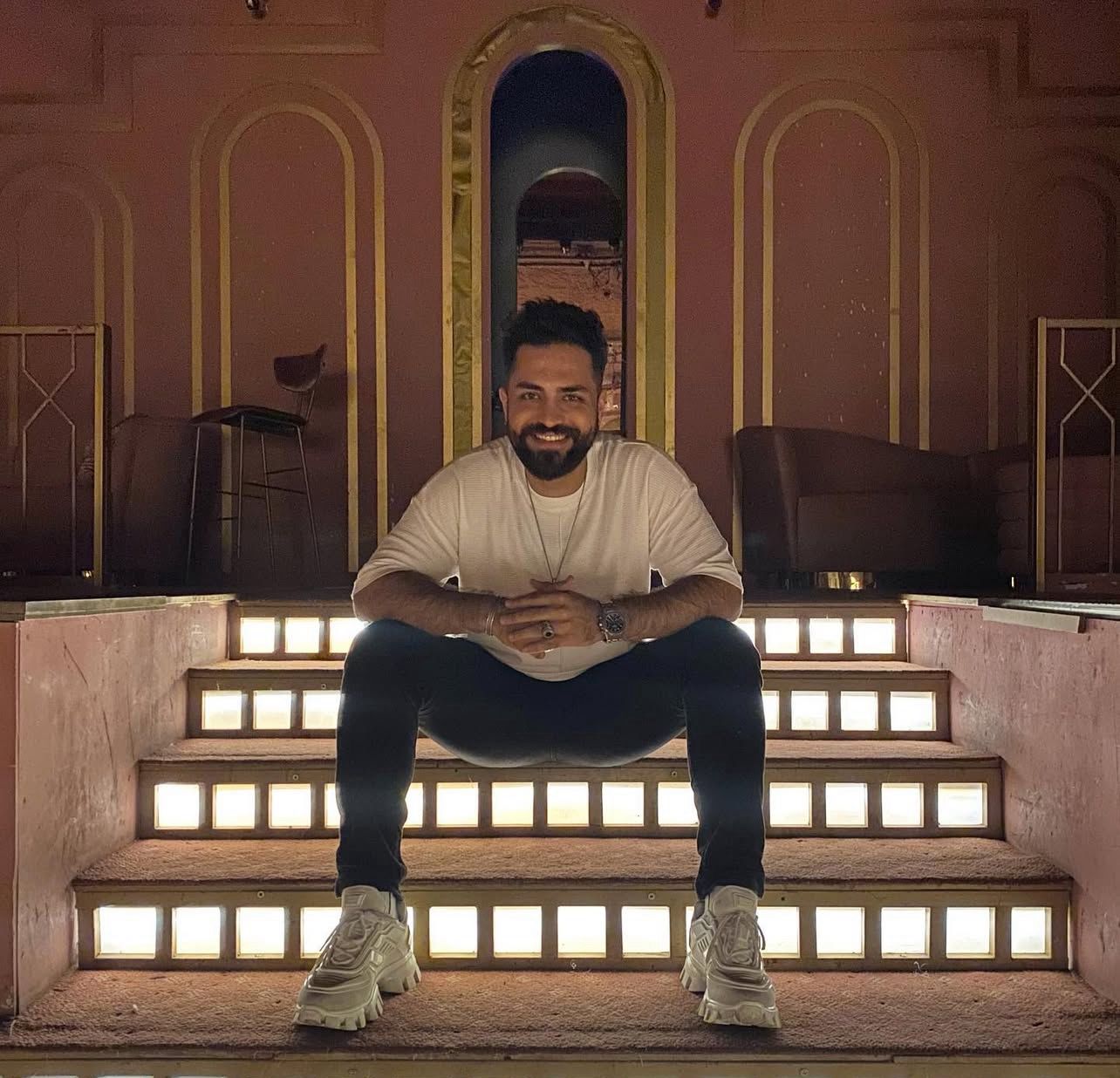 Blonde woman in a black long-sleeve and apron, arms crossed—minimalist studio portrait on a beige background.