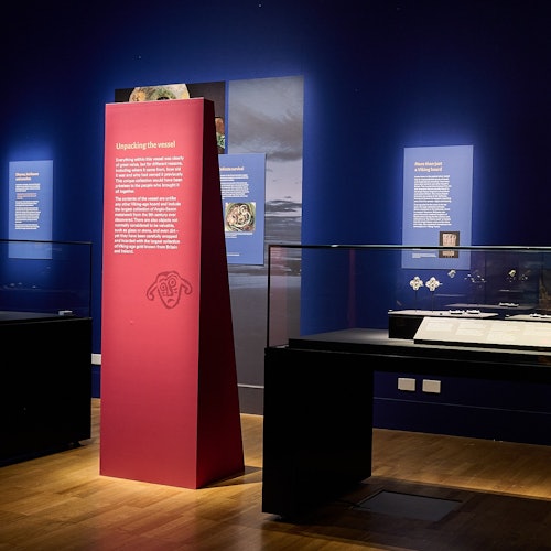 Museum exhibition room with informational display boards on dark blue walls, glass cases, and a central red information pillar.