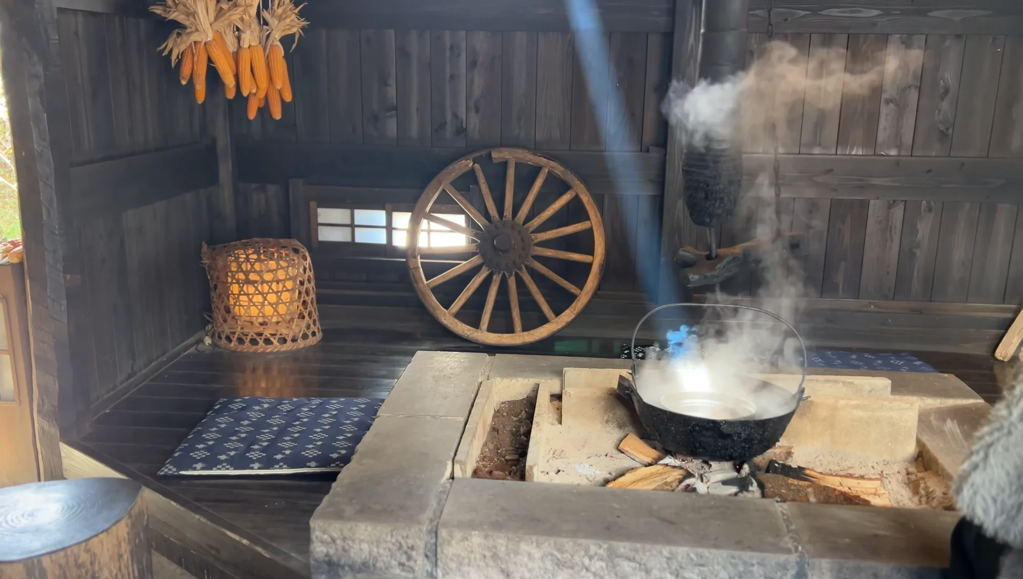 A dim old kitchen with a cauldron steaming over a stone hearth and woven baskets on the wall