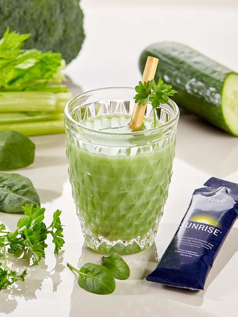 A crystal glass filled with vibrant green smoothie, garnished with fresh parsley and accompanied by leafy greens, celery, cucumber, and a wellness supplement sachet Kyäni Sunrise on a bright reflective surface.