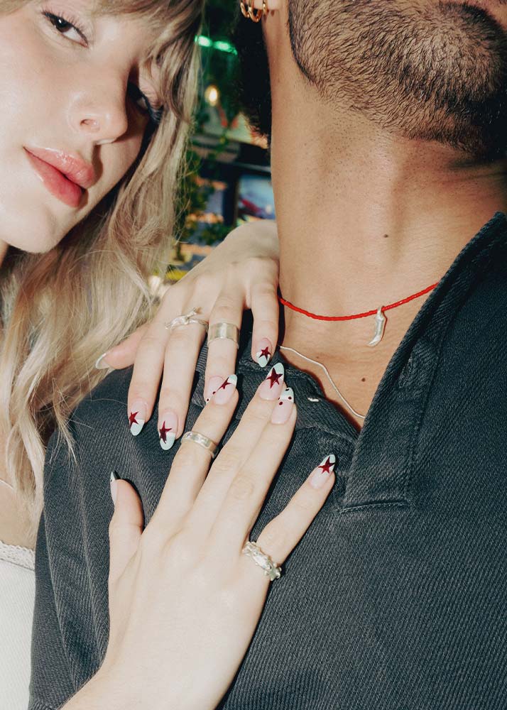 Close-up lifestyle shot of a couple with matching gothic nail art. The nails are white with red star details and black script, shown on a woman's hand resting on a man's chest, both wearing silver jewelry.
