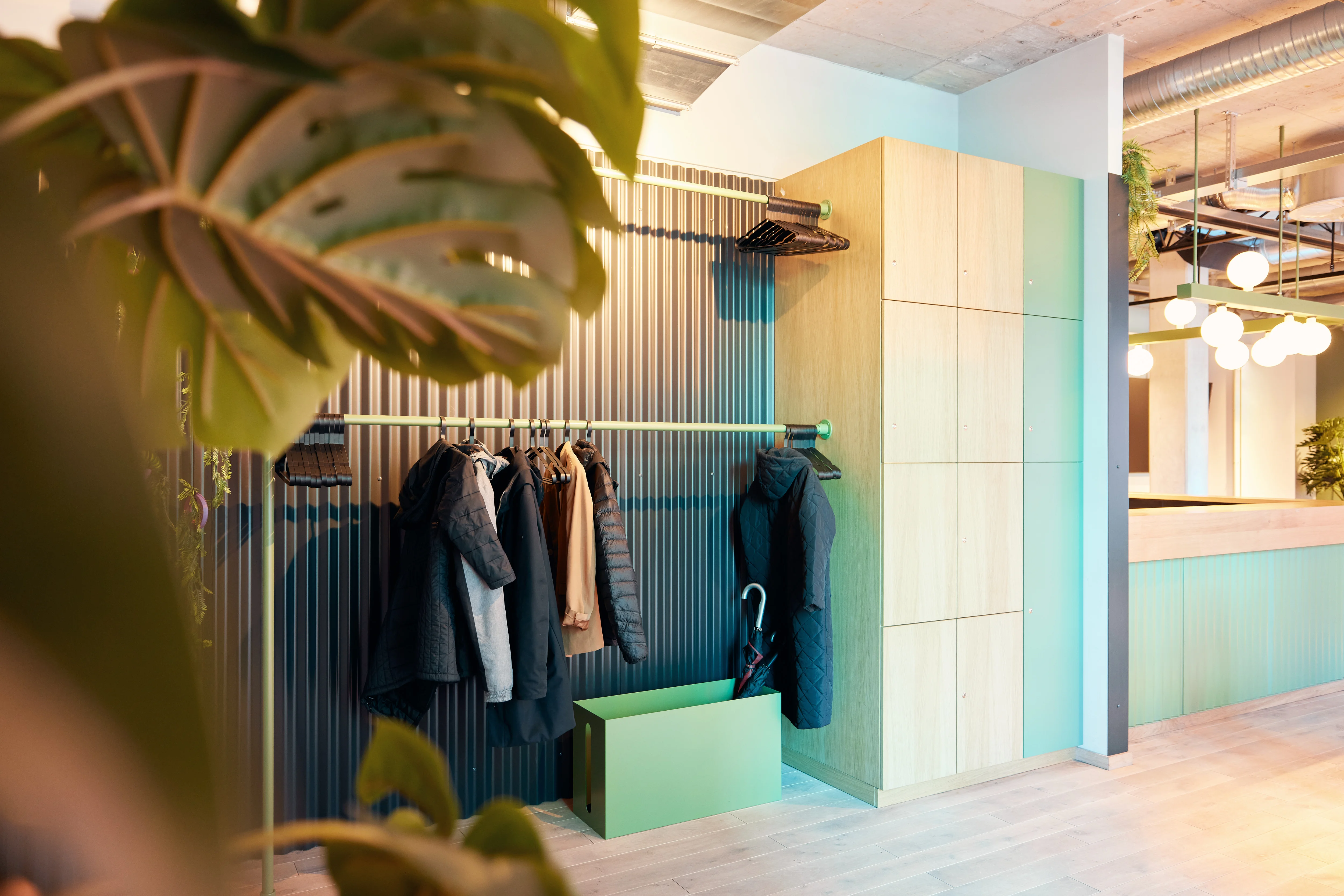 Cloakroom at 381 EVENT SPACE with coats on green rails against dark corrugated walls, wooden lockers and a green umbrella box, framed by a monstera plant.