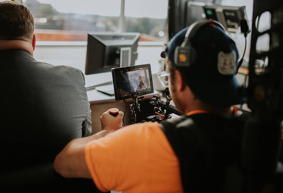A person operates a camera rig focused on a man in a suit sitting at a desk with computer monitors in an office setting.