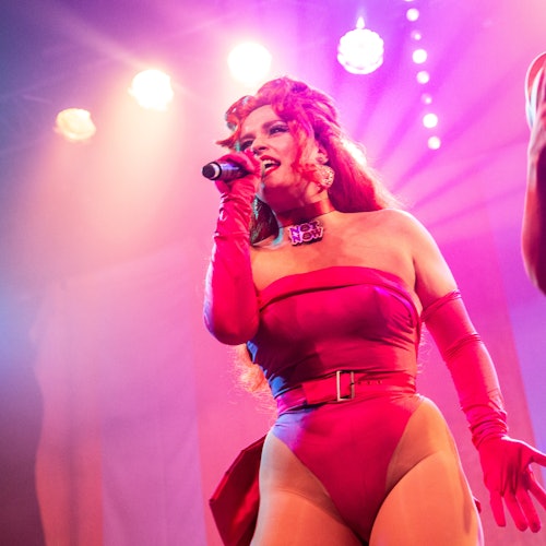 一位身穿红色服装的表演者在明亮的舞台上对着麦克风唱歌，背景是五彩斑斓的灯光。