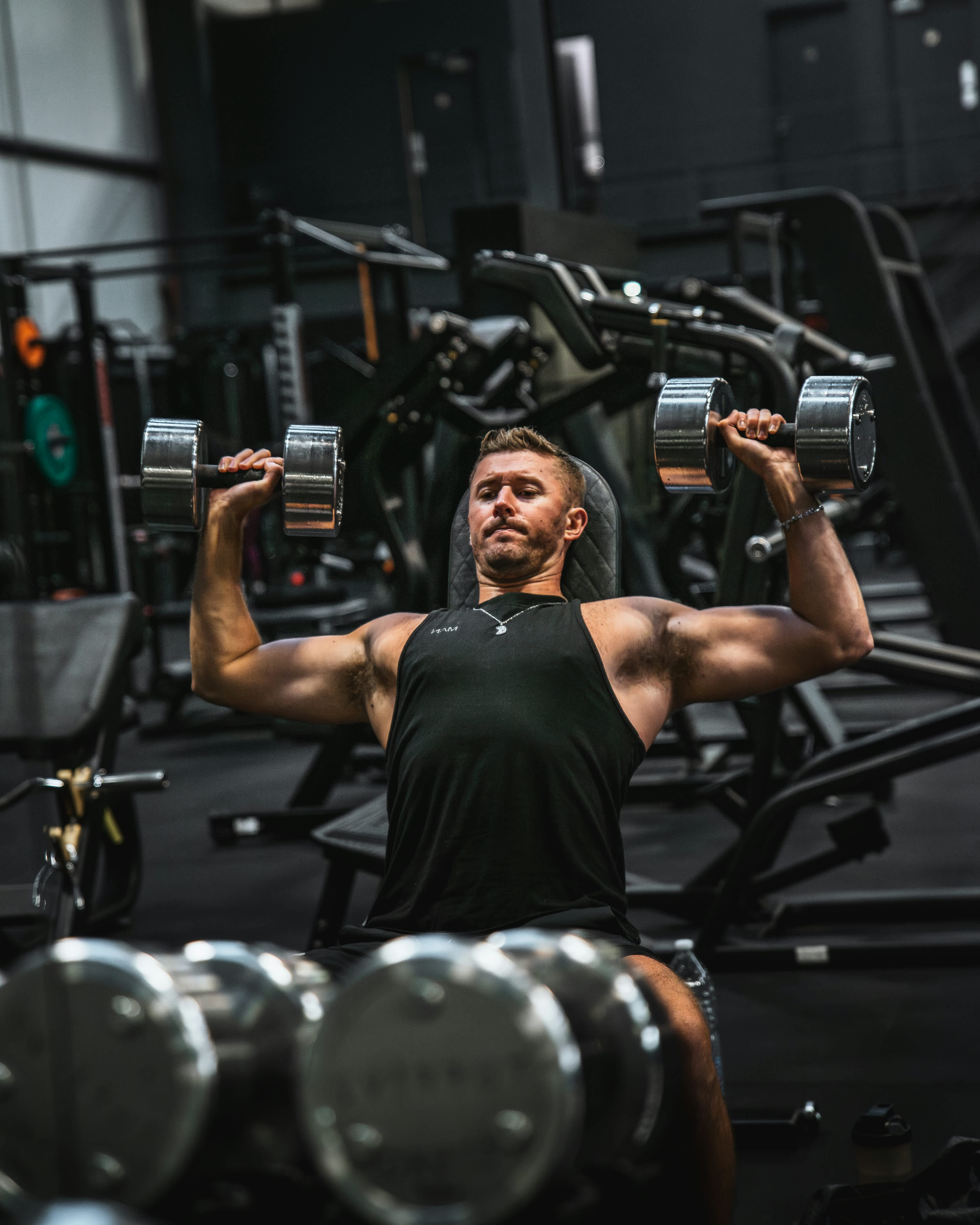 Muscular man performing a seated dumbbell shoulder press in a dark, modern gym with weight machines behind him.