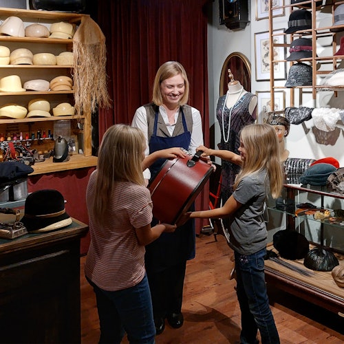 Dua anak dan seorang wanita tersenyum berada di toko topi. Anak-anak itu sedang memegang kotak topi bersama-sama. Rak menampilkan berbagai jenis topi.