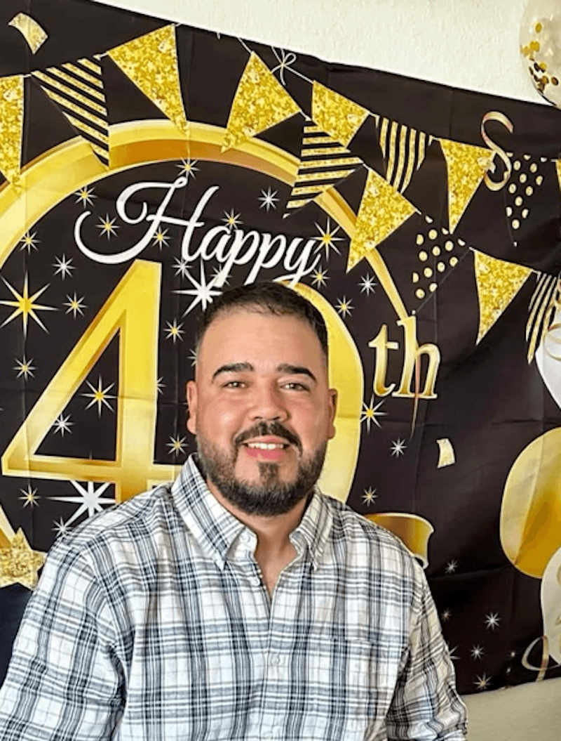 Smiling man in plaid shirt in front of a festive black and gold "Happy 40th" birthday banner with stars and bunting, creating a joyful atmosphere.