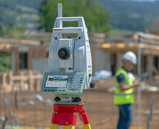 Two construction workers in safety vests and helmets review blueprints at a building site. The structured frame is visible in the background at sunset.