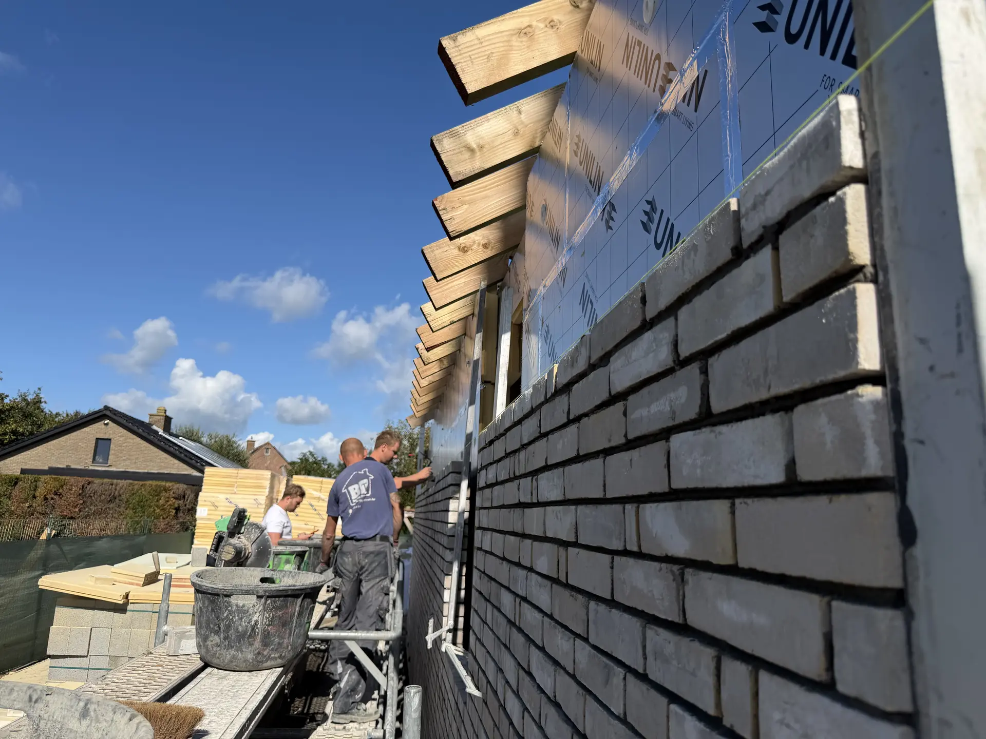 Mensen werken op een werf aan een gemetselde gevel; isolatiemateriaal en een houten constructie zijn zichtbaar.