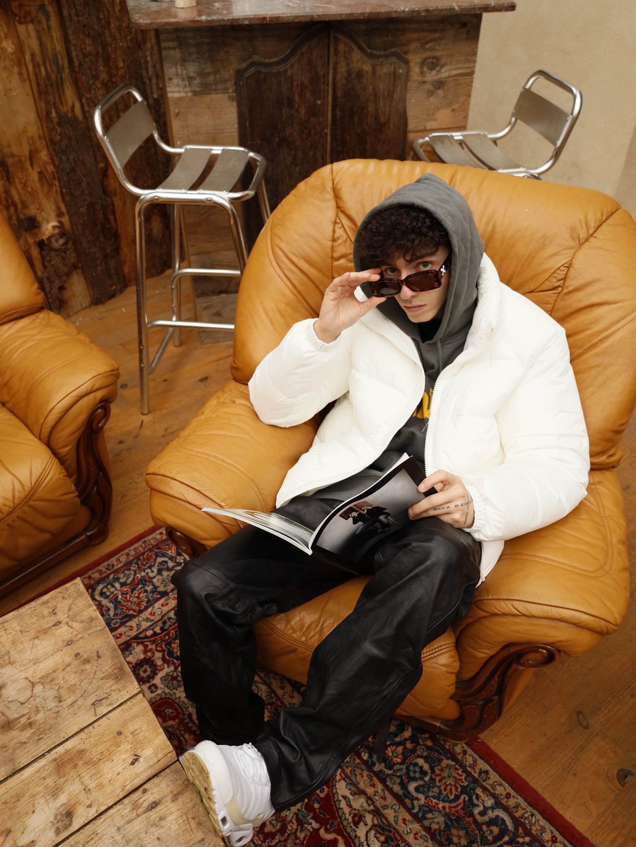 Person sitting in a cozy chair, wearing a hoodie and white jacket, looking at a magazine in a warm indoor setting.