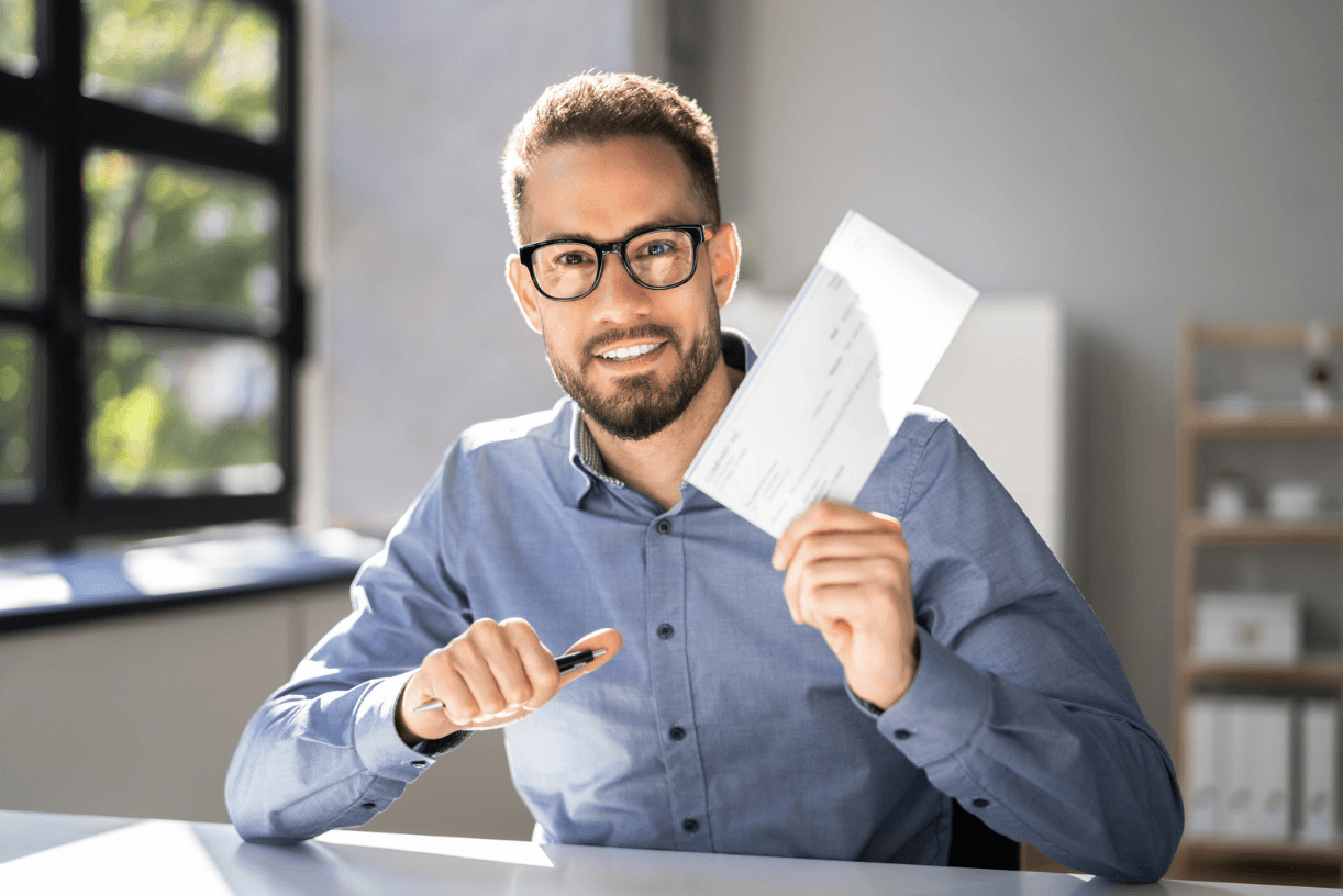 A smiling man in a blue shirt, wearing glasses, and holding a piece of paper in his hand. He appears to be sitting at a desk in an office or work environment.