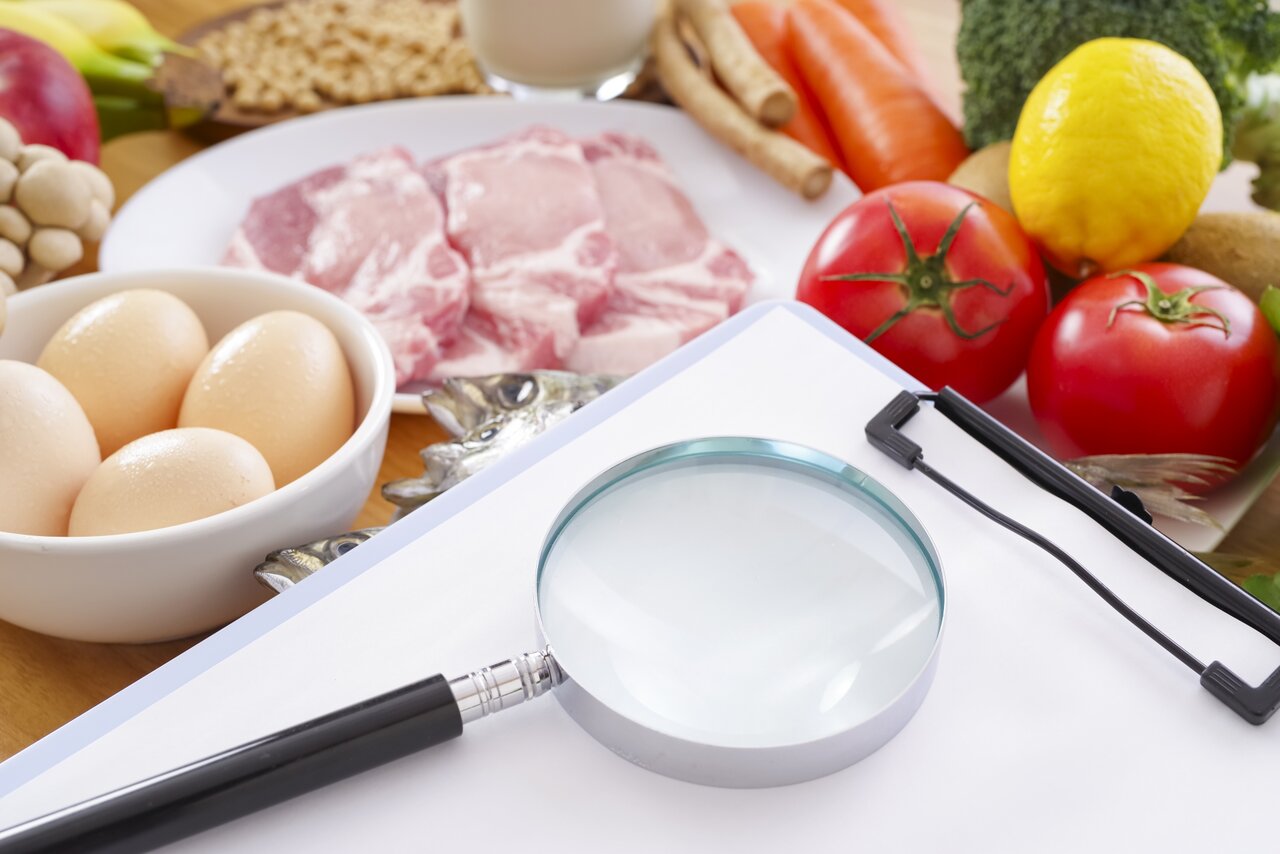 食材の横に置かれた虫めがねと白いボード。成分表示やエビデンスをWeb上に検証可能な形で置き、広告表現やパッケージ表示との食い違いをなくす準備を表す