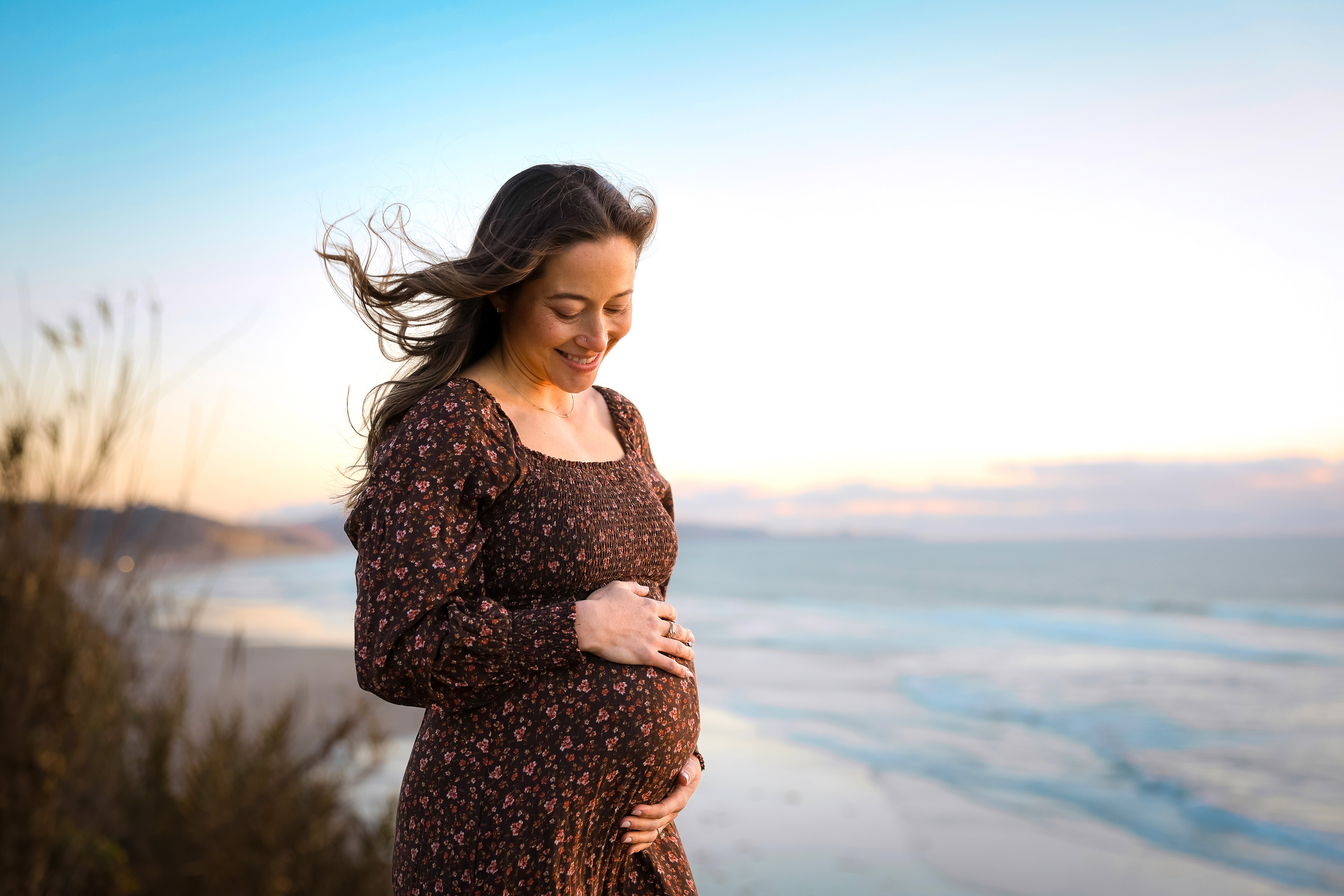 Pregnant woman standing by the ocean at sunset, holding her belly during a maternity session in San Diego.