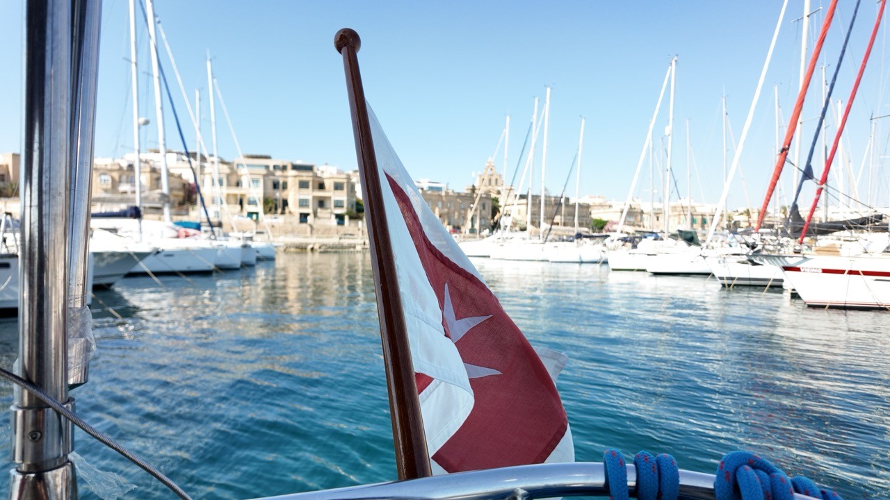 Close-up view from the stern of a sailboat showing a Maltese maritime ensign — a red and white flag bearing a white Maltese cross — flying from the backstay. A busy marina filled with white-hulled sailboats and traditional Maltese stone architecture is softly blurred in the background.